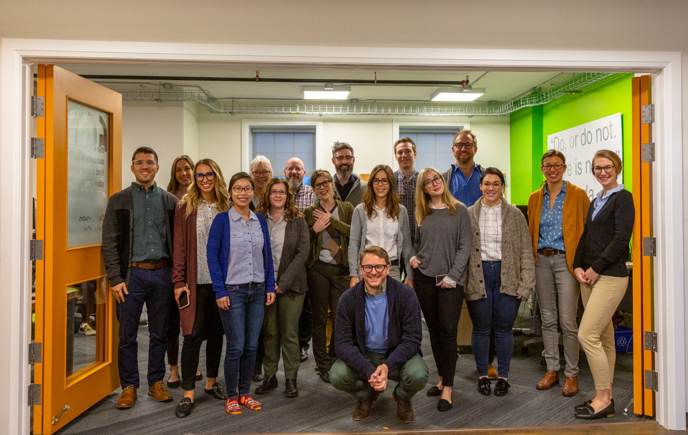 A group of thirteen people is standing and one person is crouching inside an office with green walls and an open orange door.