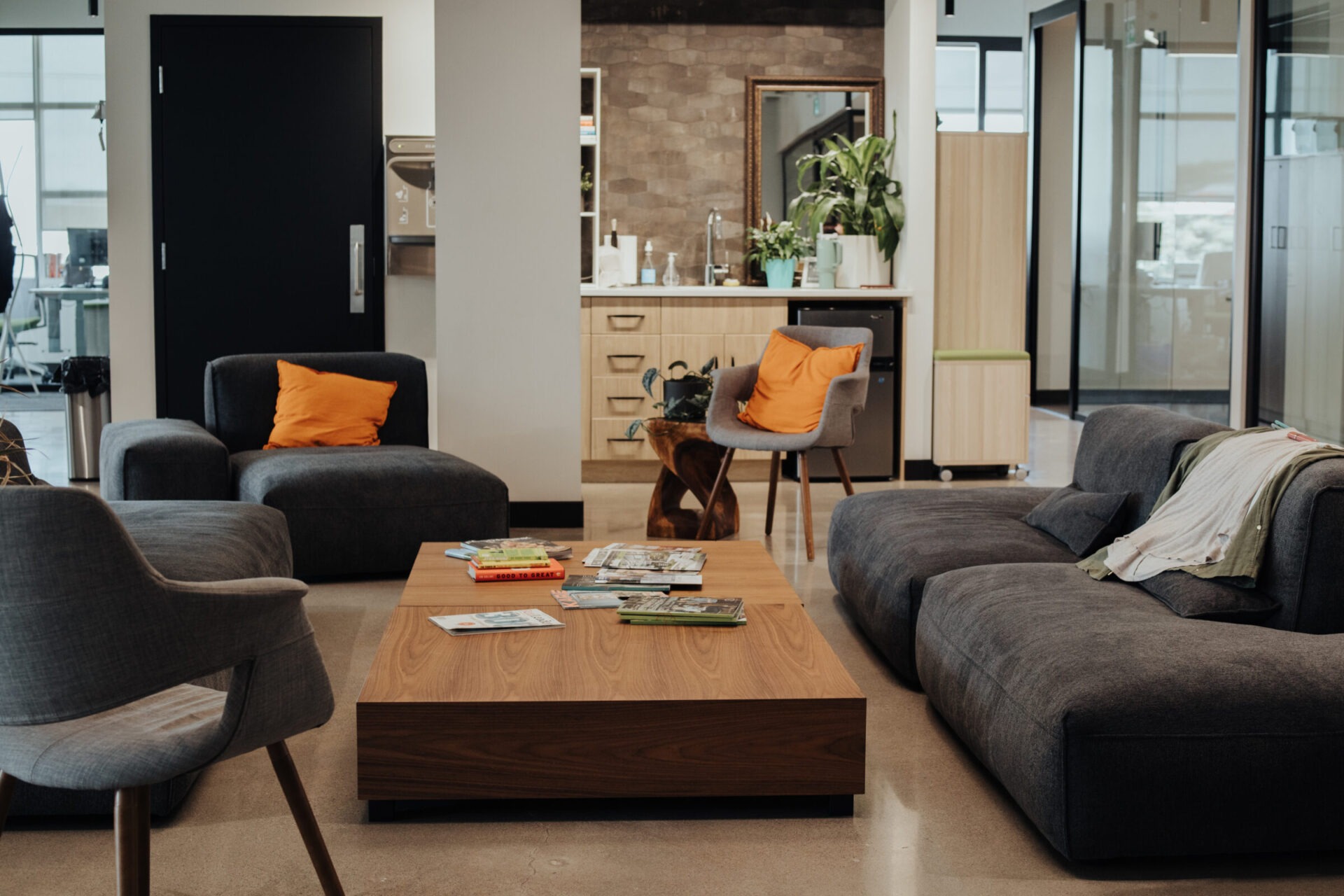 A modern, cozy office lounge with grey sofas, orange pillows, a wooden coffee table, magazines, indoor plants, and a kitchenette in the background.