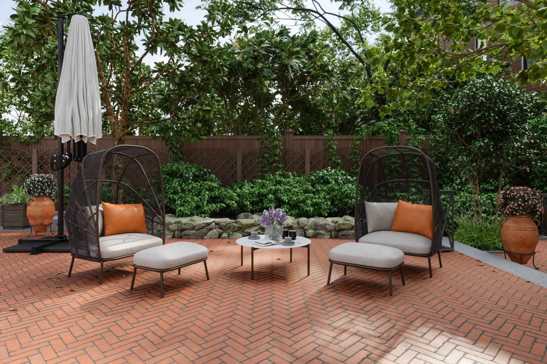 A tranquil patio with wicker chairs, footrests, a white table, umbrella, terracotta planters, and leafy greenery in the background. No people are present.