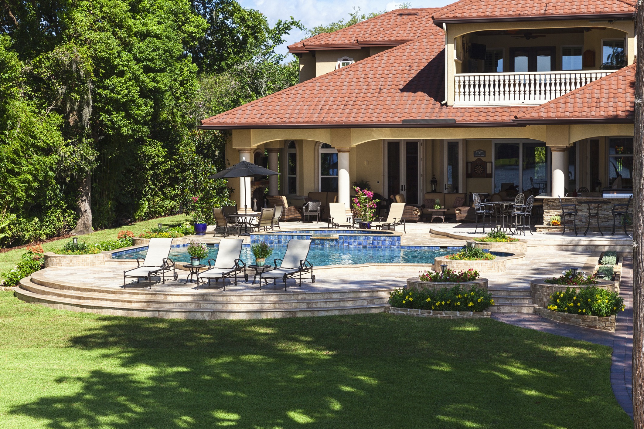 A luxurious house with a red-tiled roof, pool, lounge chairs, and lush greenery surrounding a spacious backyard.