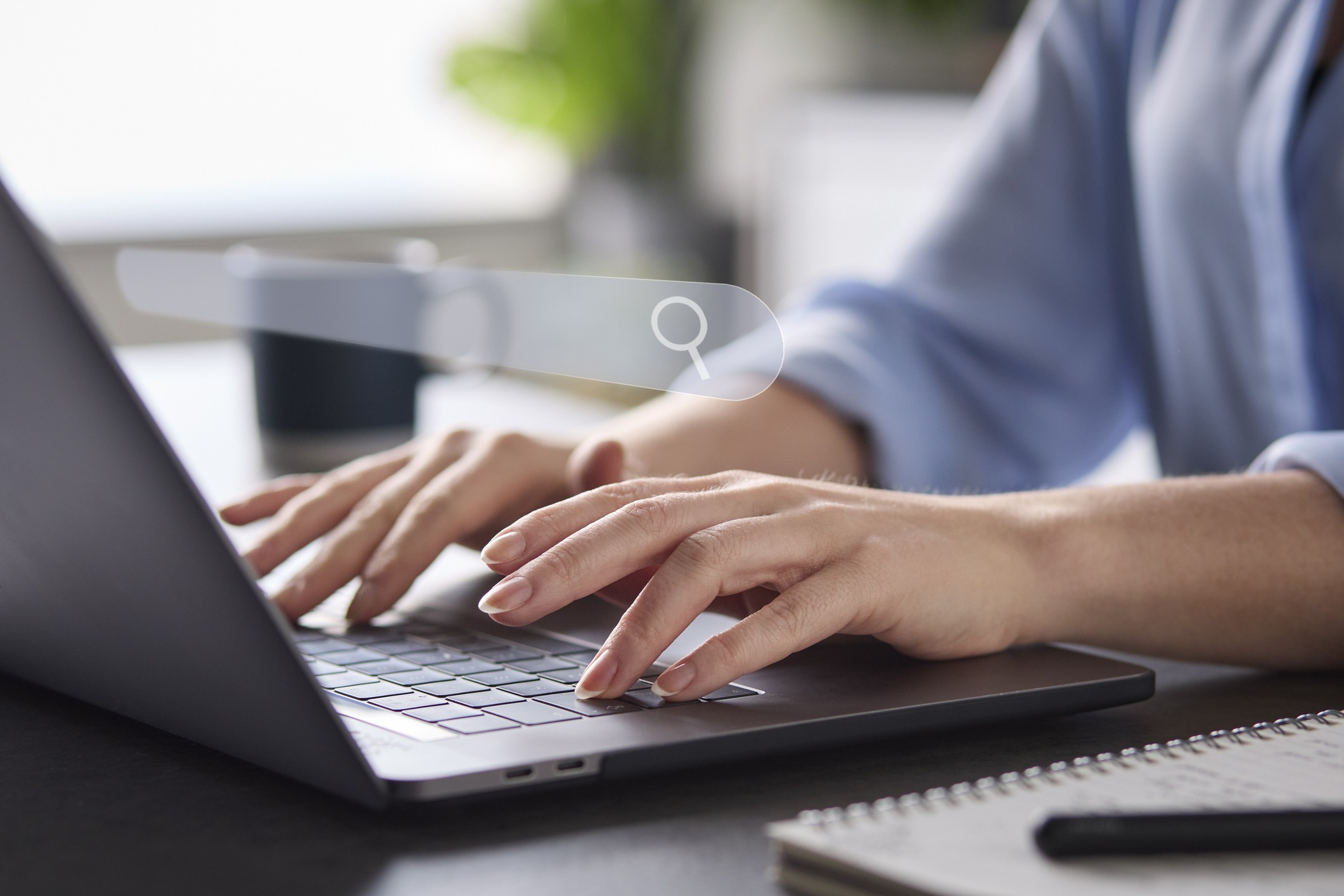 A person typing on a laptop with a search icon overlay, next to a notebook and pen in a bright, modern workspace.