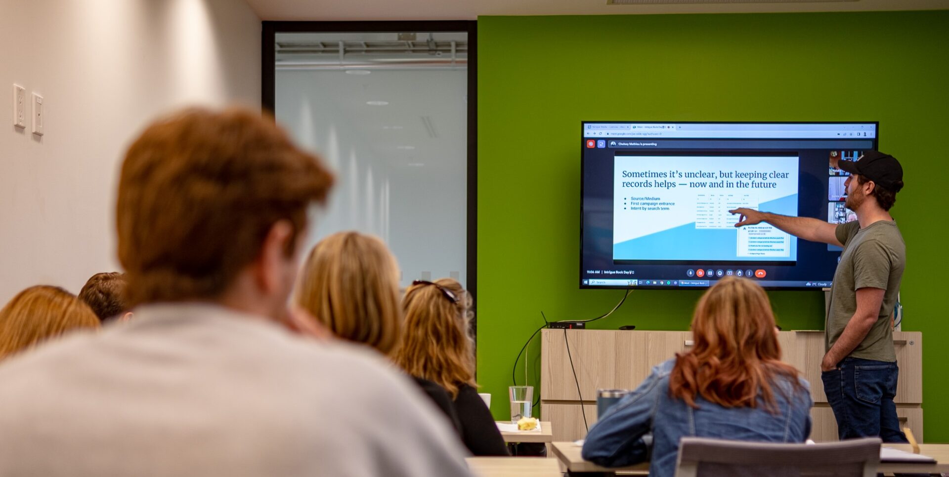 A person presents a detailed slide to a focused audience in a modern classroom setting. The background features a green wall.