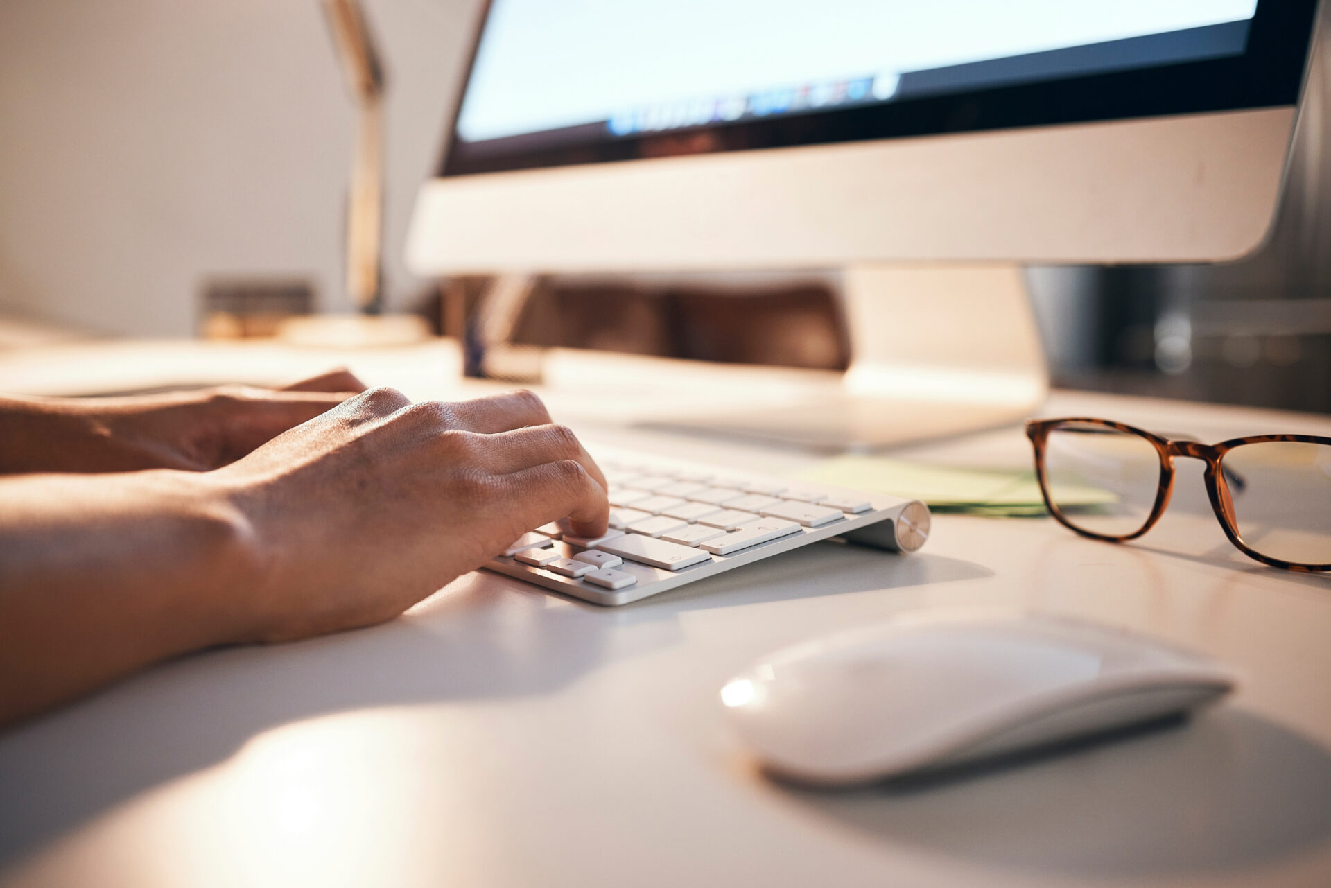 A person types on a computer keyboard next to a mouse and eyeglasses, all situated on a clean, modern desk.