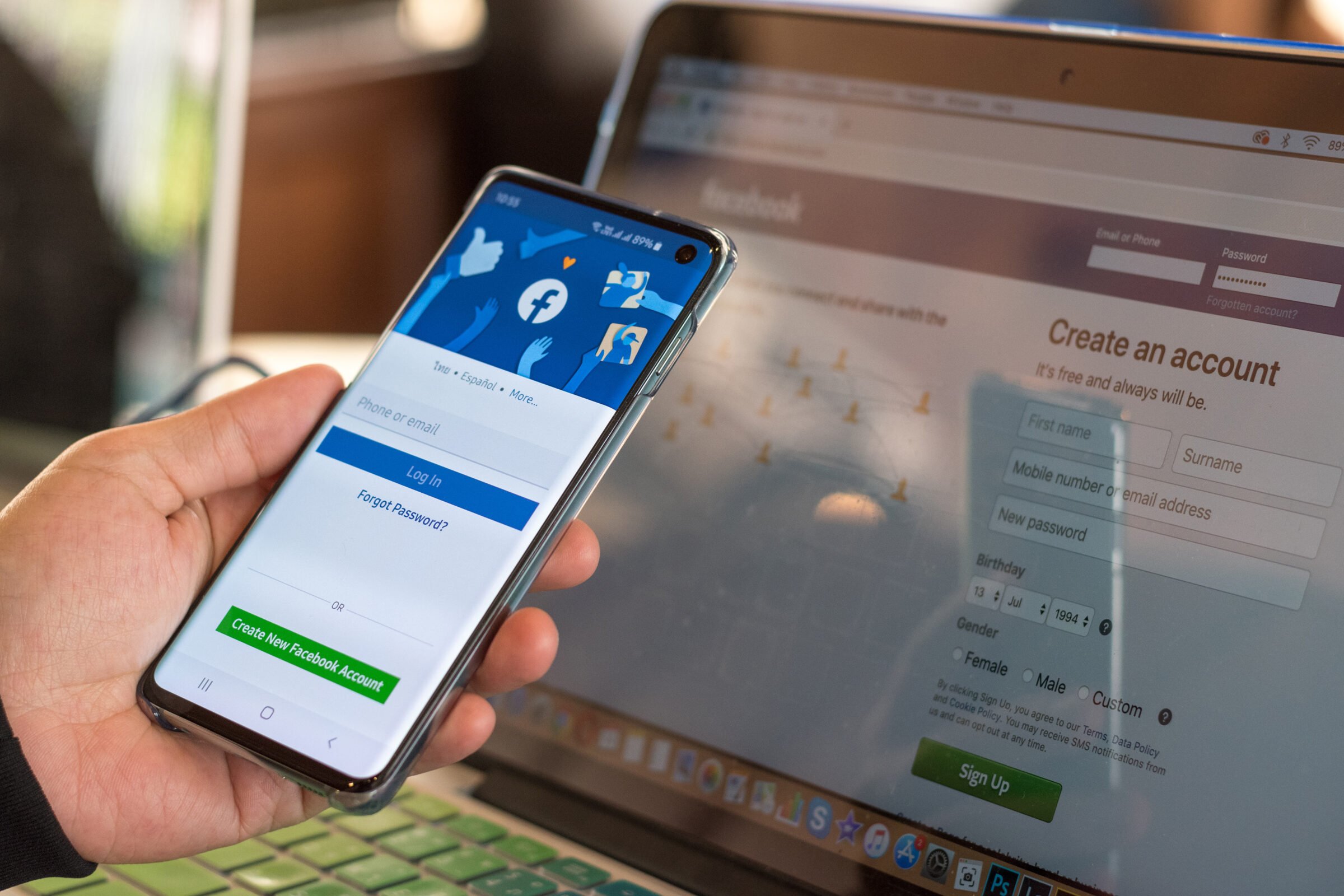 A person is holding a smartphone with Facebook's login screen, next to a laptop displaying Facebook's account creation page.