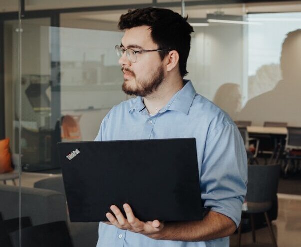 A person holding a laptop stands in a modern office with glass walls, illuminated by geometric ceiling lights.