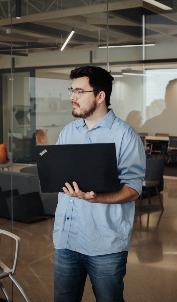 A person holding a laptop stands in a modern office with glass walls, illuminated by geometric ceiling lights.