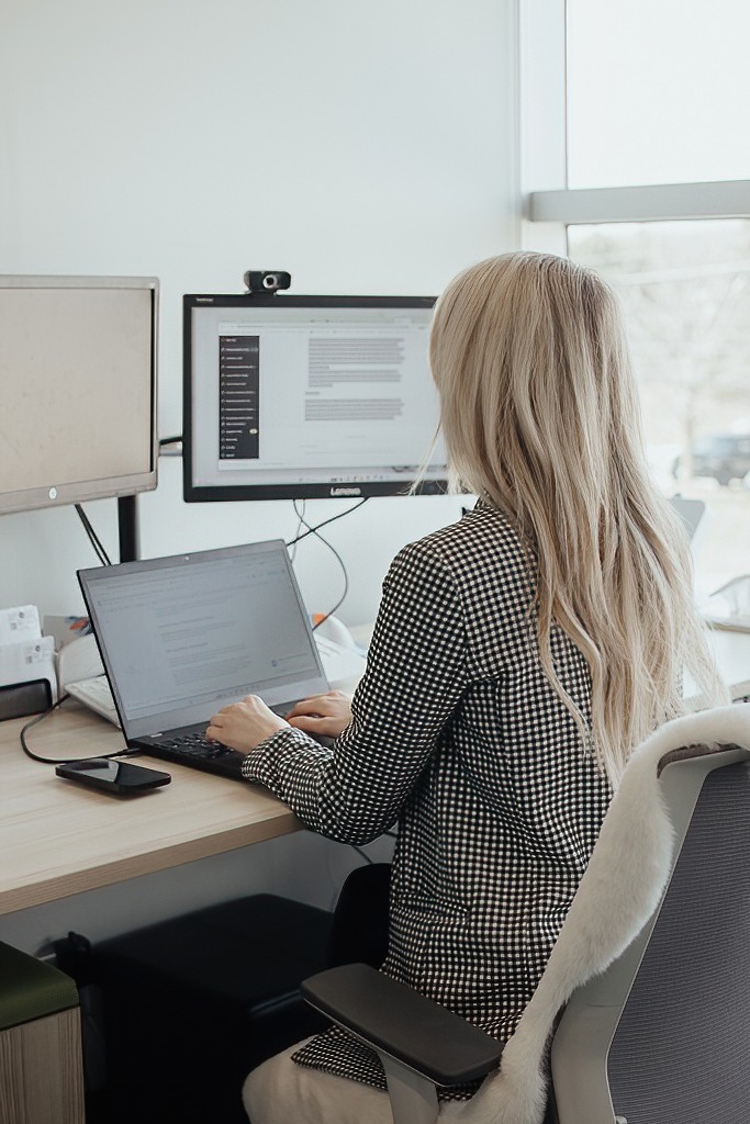 A person sits at a desk using a laptop and dual monitors in a bright office with large windows and natural light.