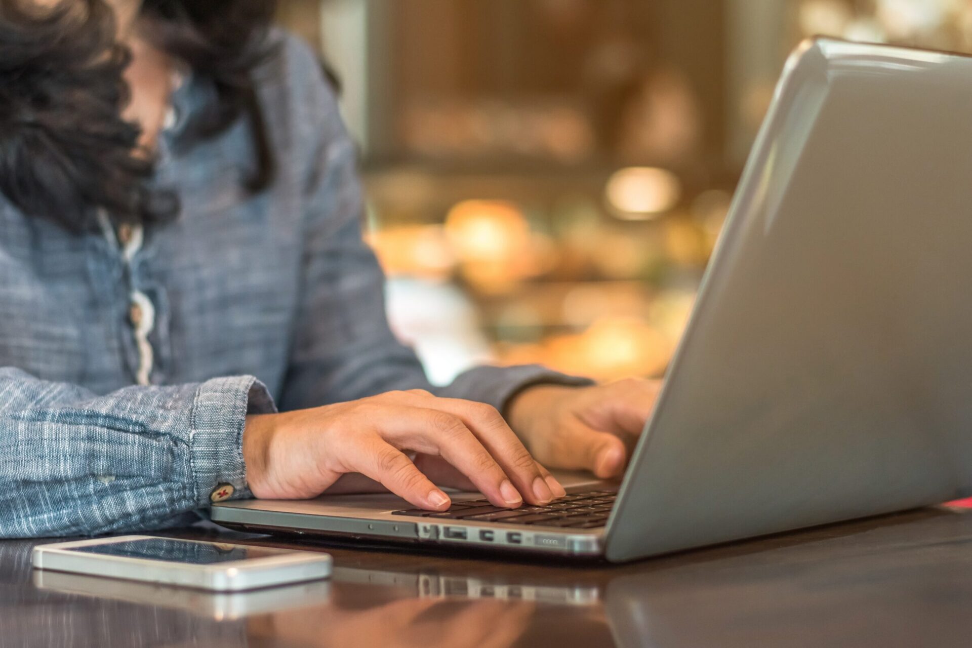 A person in casual attire types on a laptop next to a smartphone, sitting at a table in a blurred indoor setting.