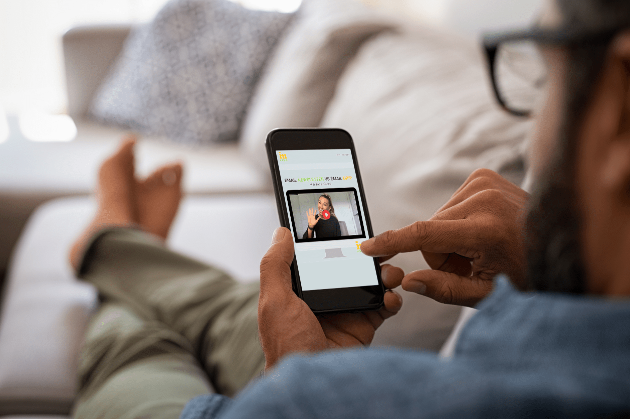 A person relaxes on a couch, viewing a video on a smartphone about email newsletters and email drip campaigns.