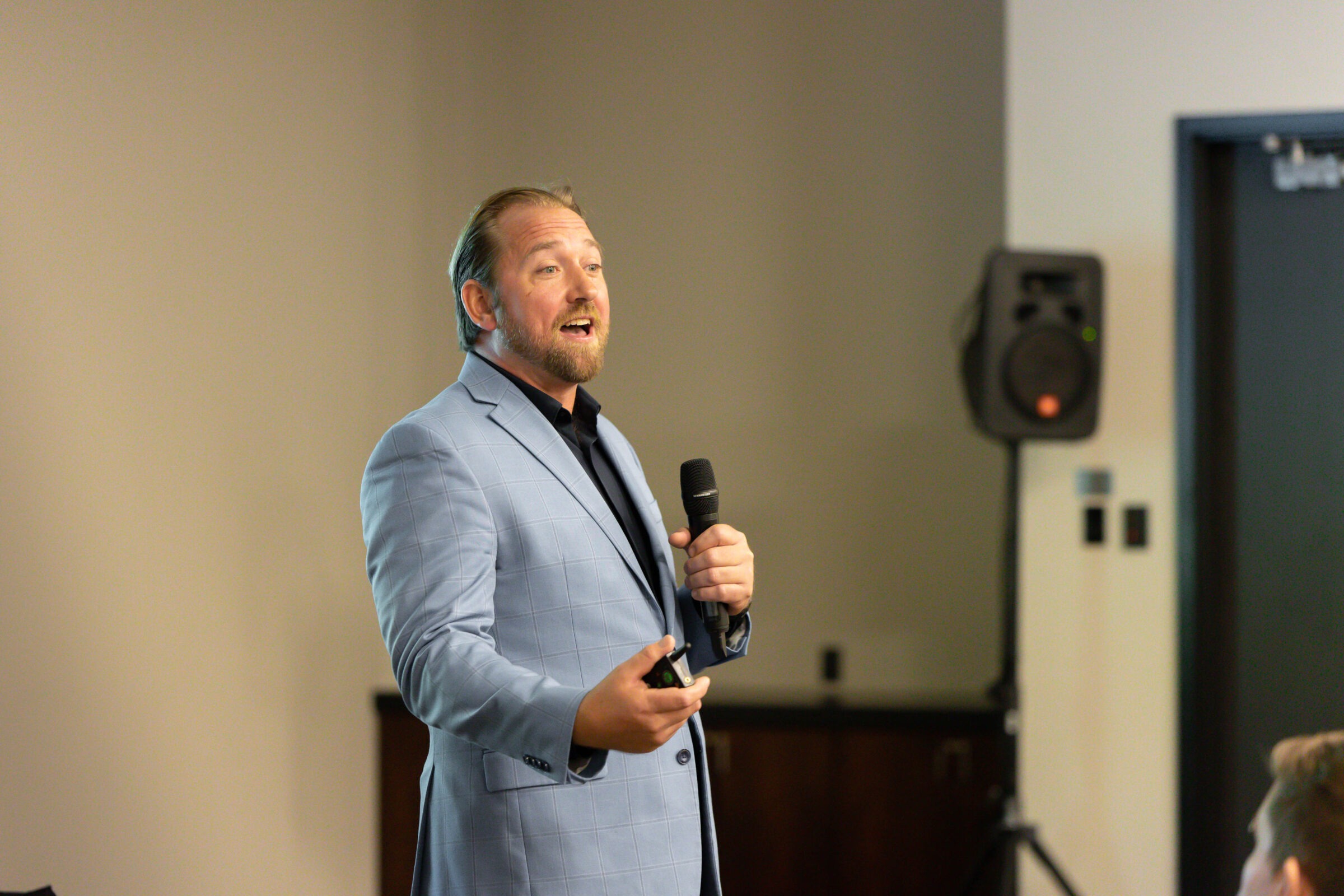 A person in a light blue blazer is giving a presentation, holding a microphone and remote, with a speaker in the background.