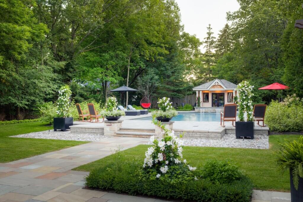 A serene backyard with a pool, lush greenery, lounge chairs, a small pavilion, and vibrant flowers on a sunny day.