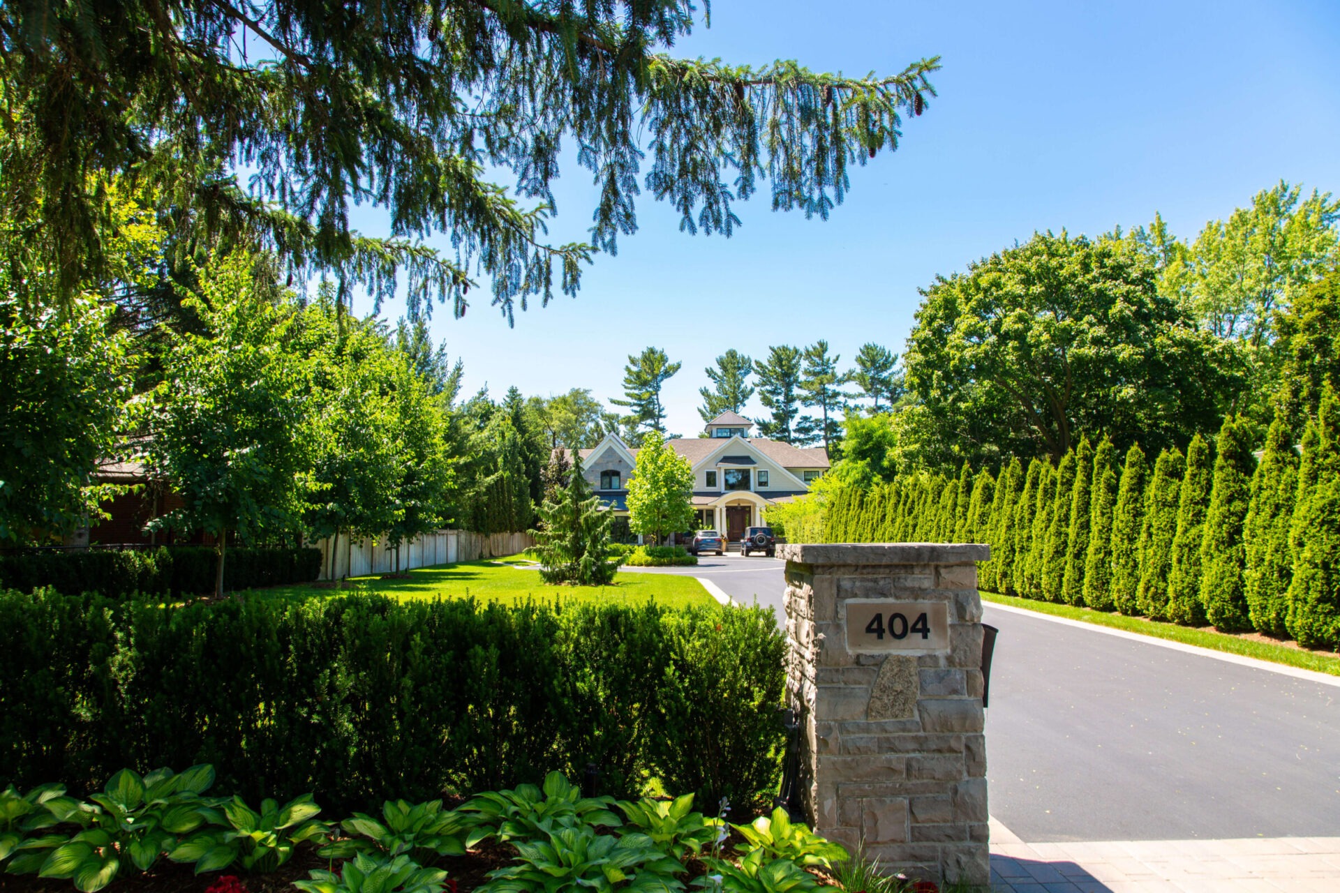 Charming house with manicured lawn and trees, viewed from driveway entrance marked "404," bordered by lush greenery and flanked by stone pillars.