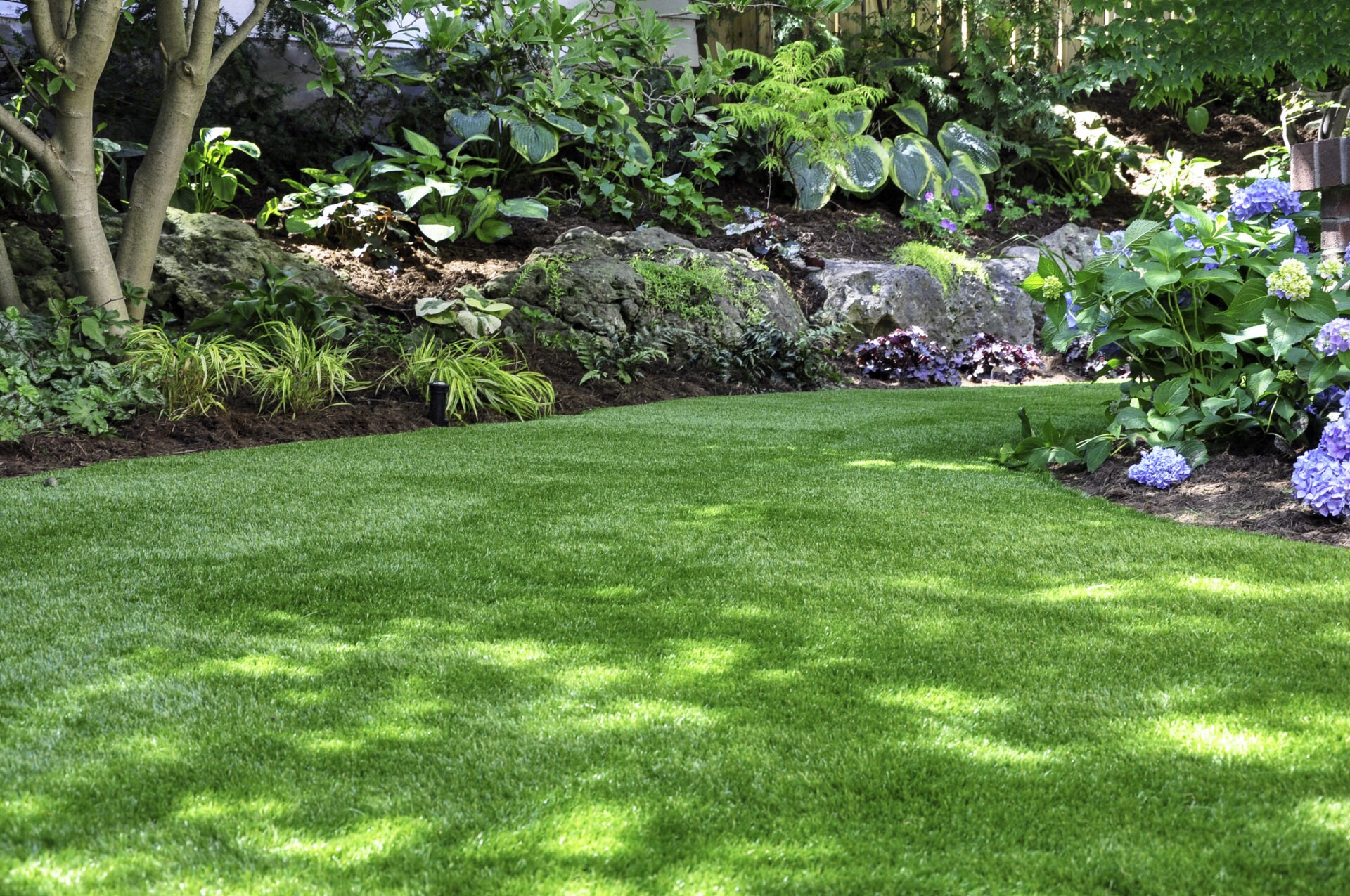 A lush garden with vibrant green grass, colorful flowers, leafy shrubs, and a border of rocks under partially shaded sunlight. No landmarks visible.