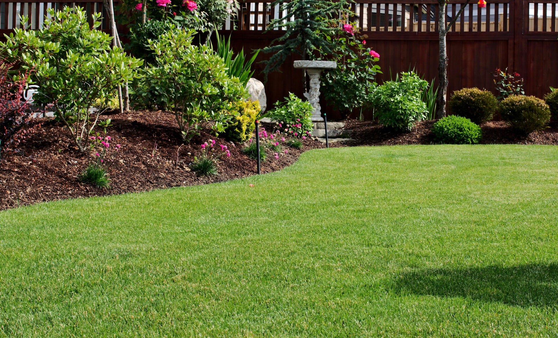 A well-manicured garden has lush green grass, trimmed bushes, vibrant flowers, and a birdbath, enclosed by a wooden fence in the background.