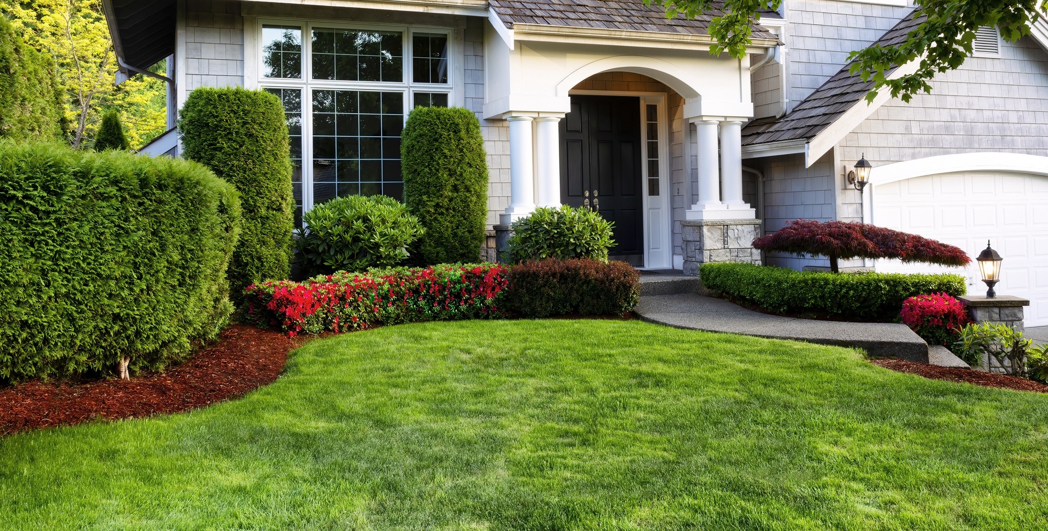 A well-manicured lawn with lush hedges and vibrant flowerbeds in front of a charming house featuring a columned entrance and large windows.