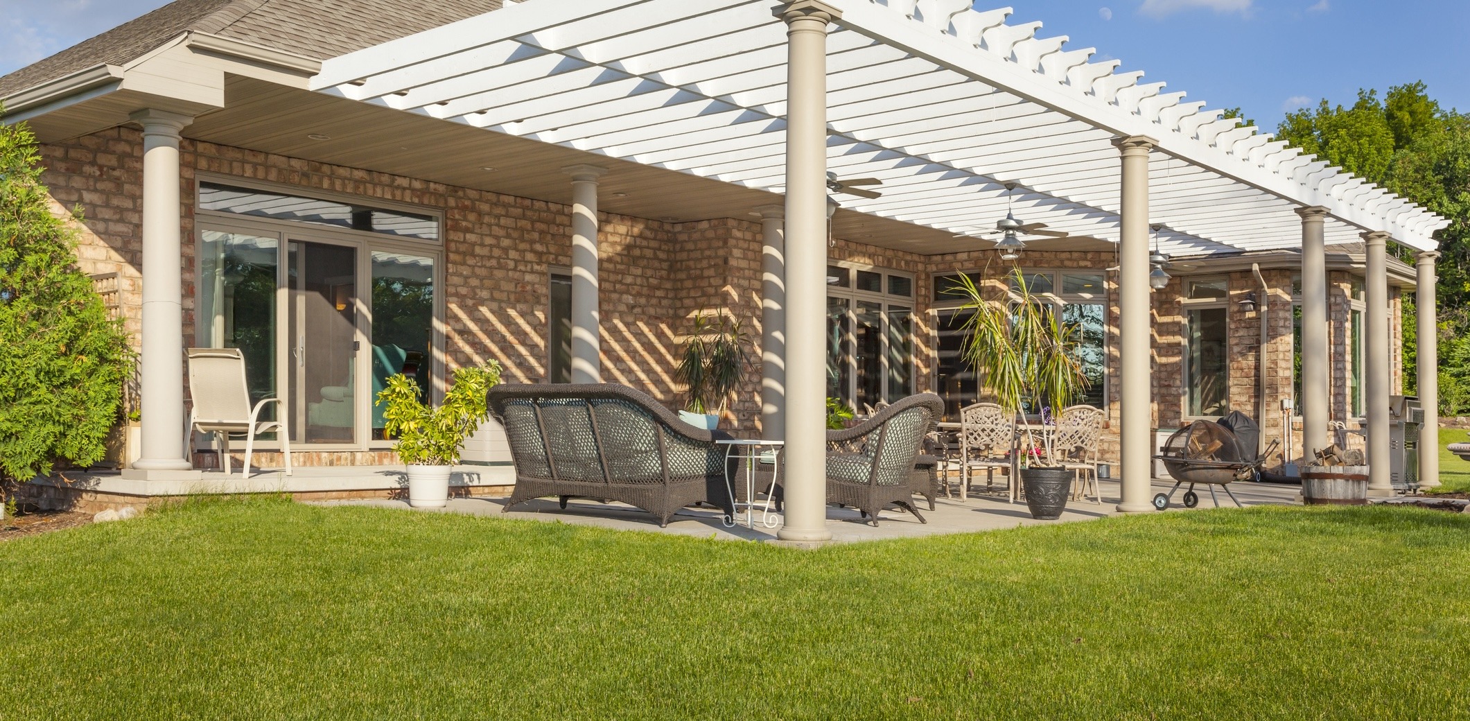 A spacious patio with wicker furniture and potted plants shaded by a pergola, adjoining a brick house with large glass doors.