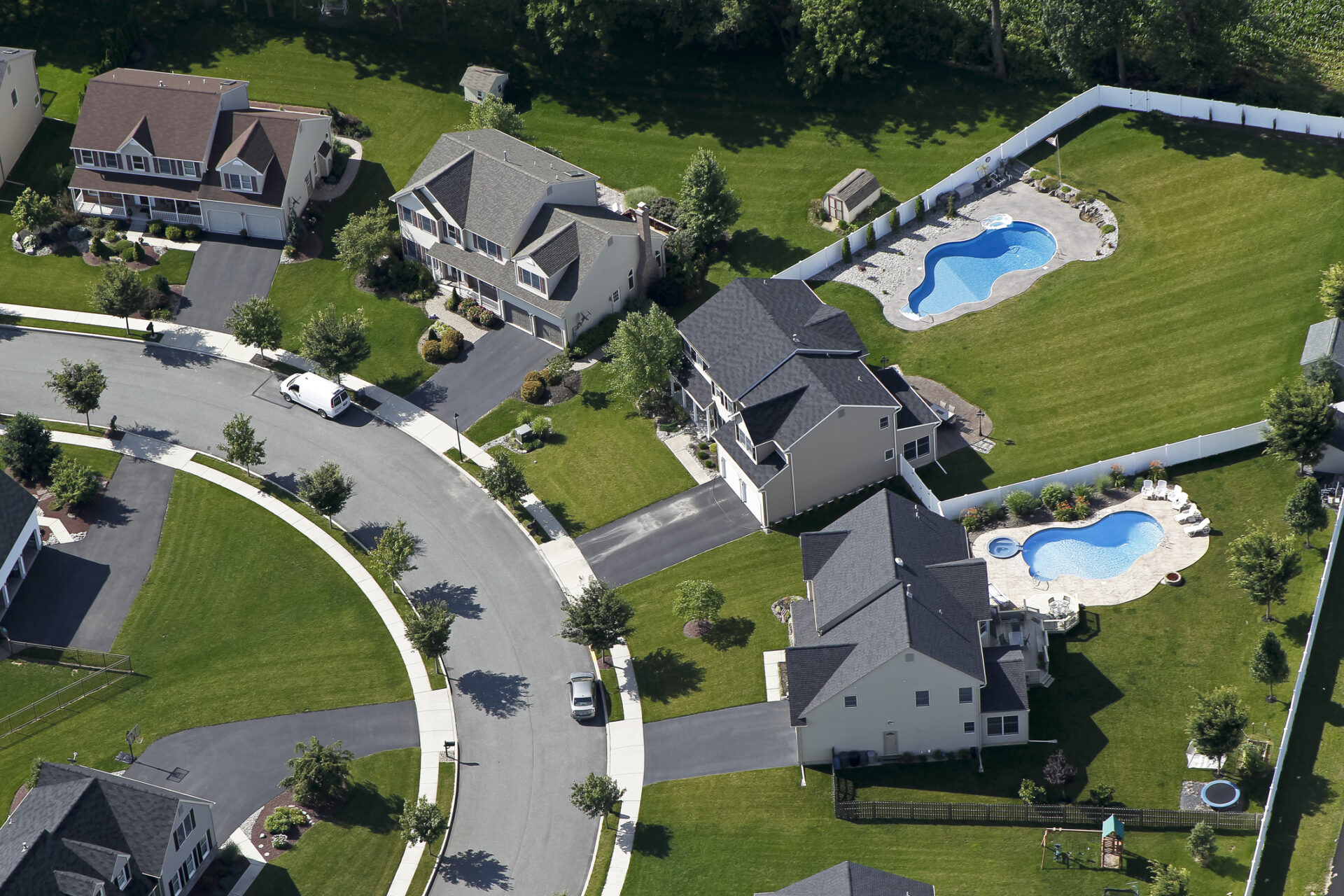 Aerial view of suburban neighborhood with several houses, backyards featuring pools, green lawns, curved street, and well-maintained landscaping. No historical buildings visible.