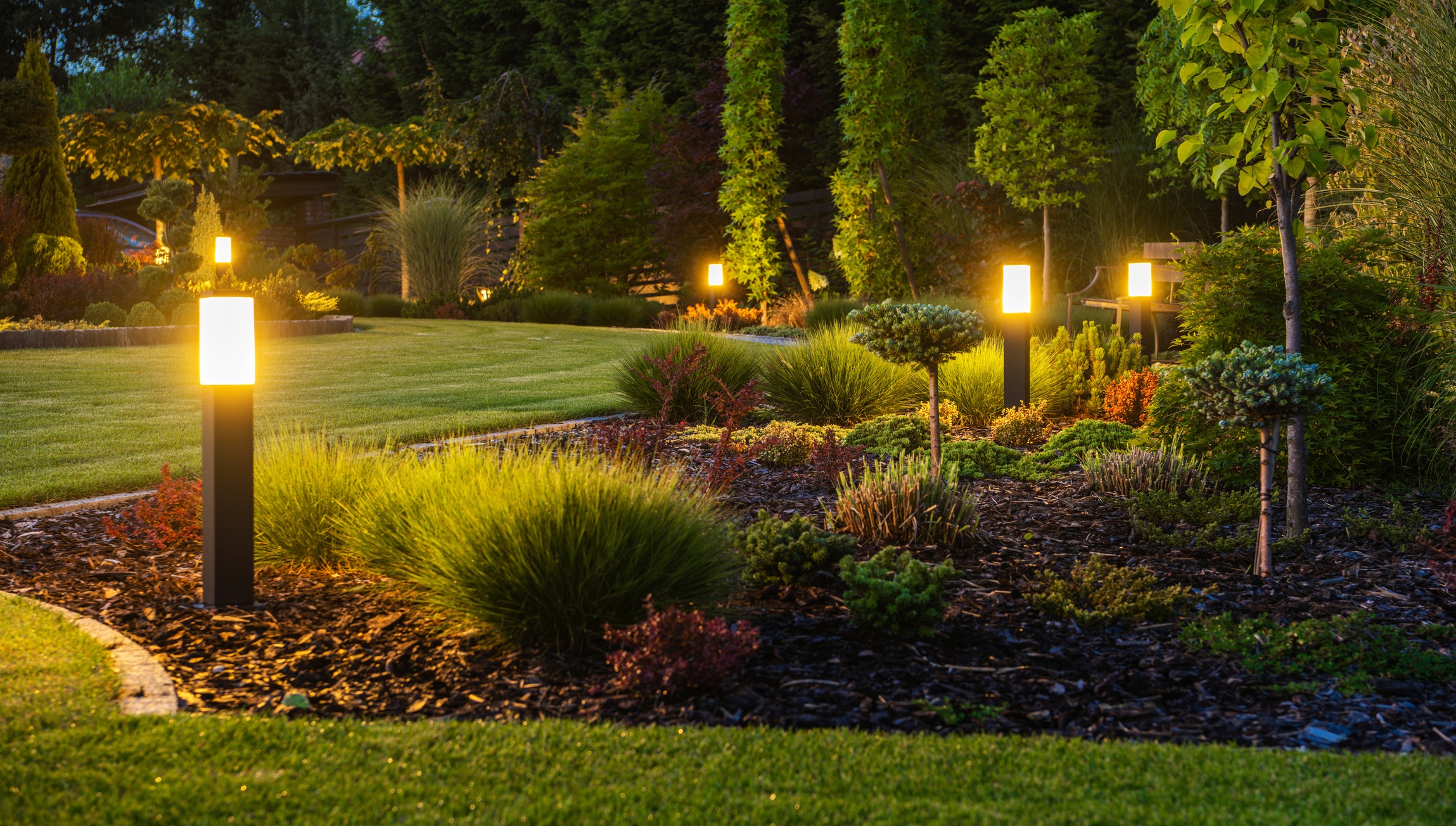 A well-maintained garden at dusk, featuring illuminated pathway lights, various shrubs, trees, and carefully landscaped greenery with a serene and inviting atmosphere.
