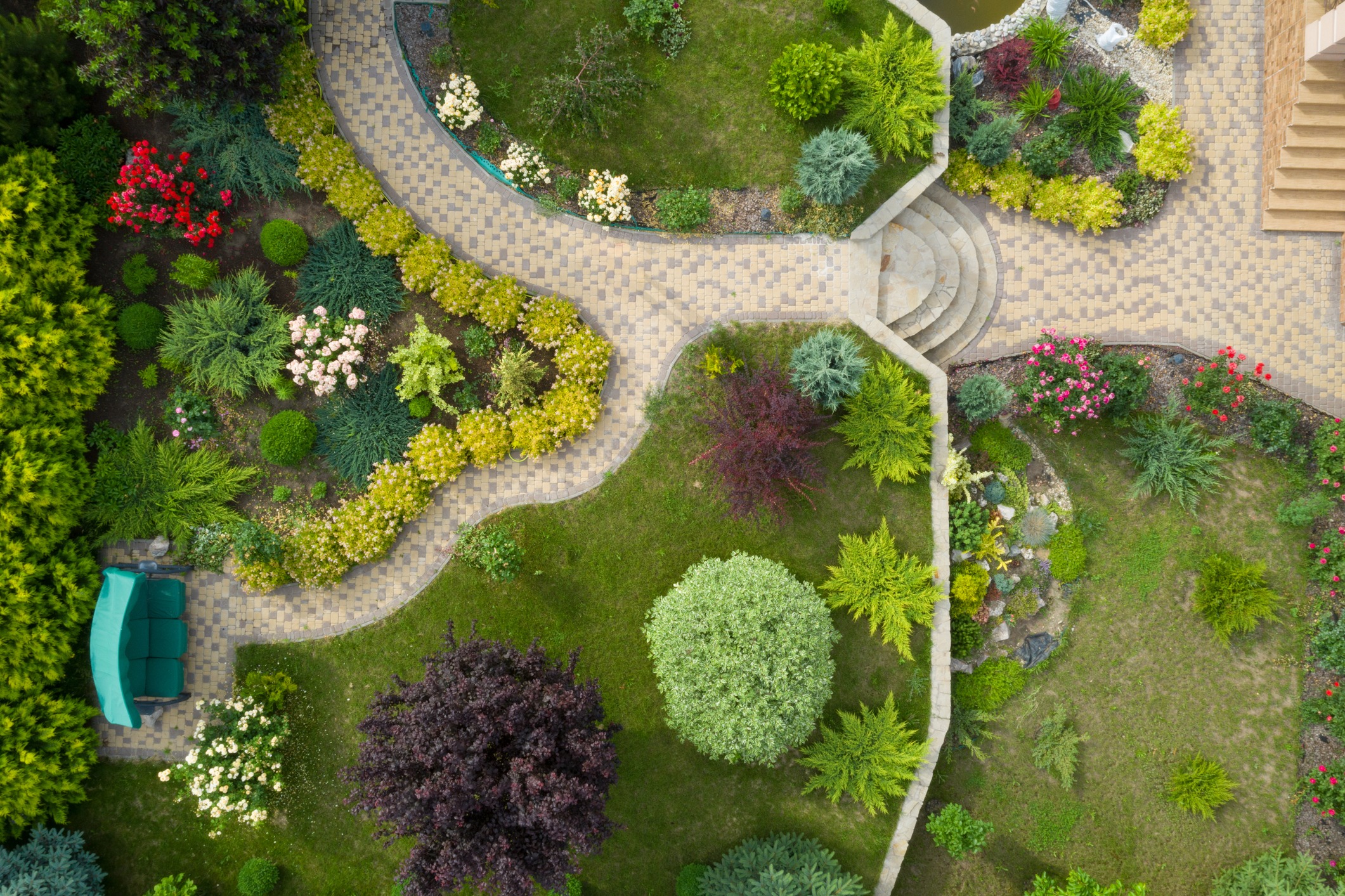 An aerial view of a garden with manicured lawns, shrubs, flowers, and curved brick walkways, featuring a green bench and stone steps.
