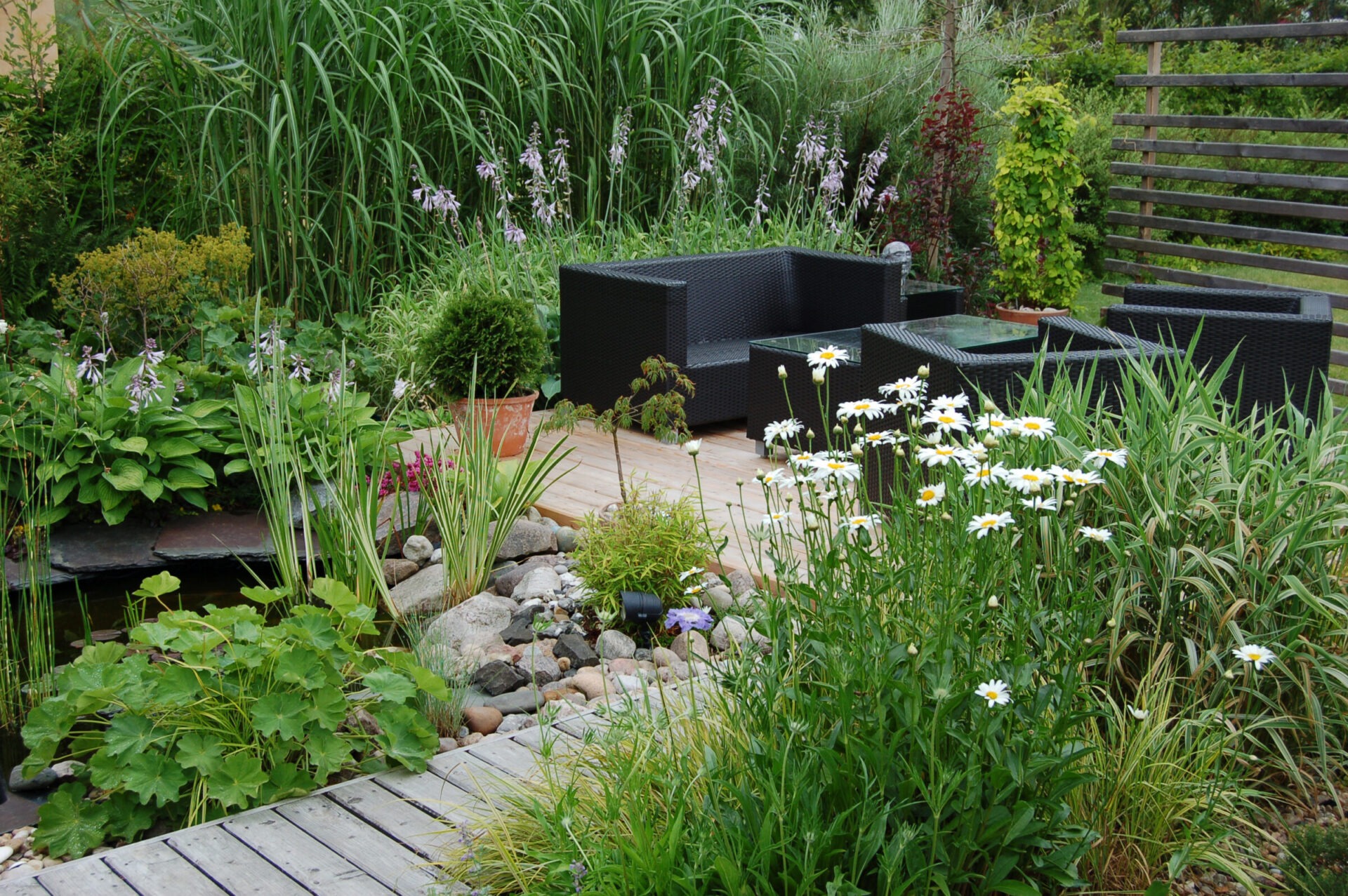 A vibrant garden with various flowers, lush greenery, wooden deck, black wicker furniture, and a small rock-bordered pond, creating a serene outdoor seating area.