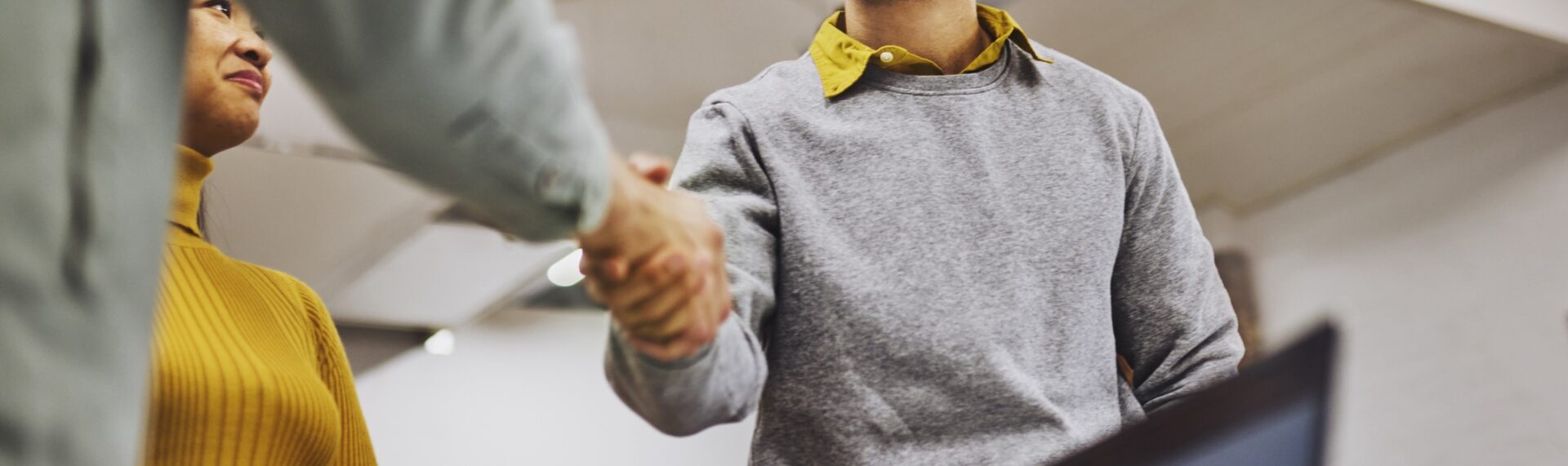 Two people are shaking hands, one wearing a mustard sweater and another in a grey sweater with a yellow collar in an indoor setting.