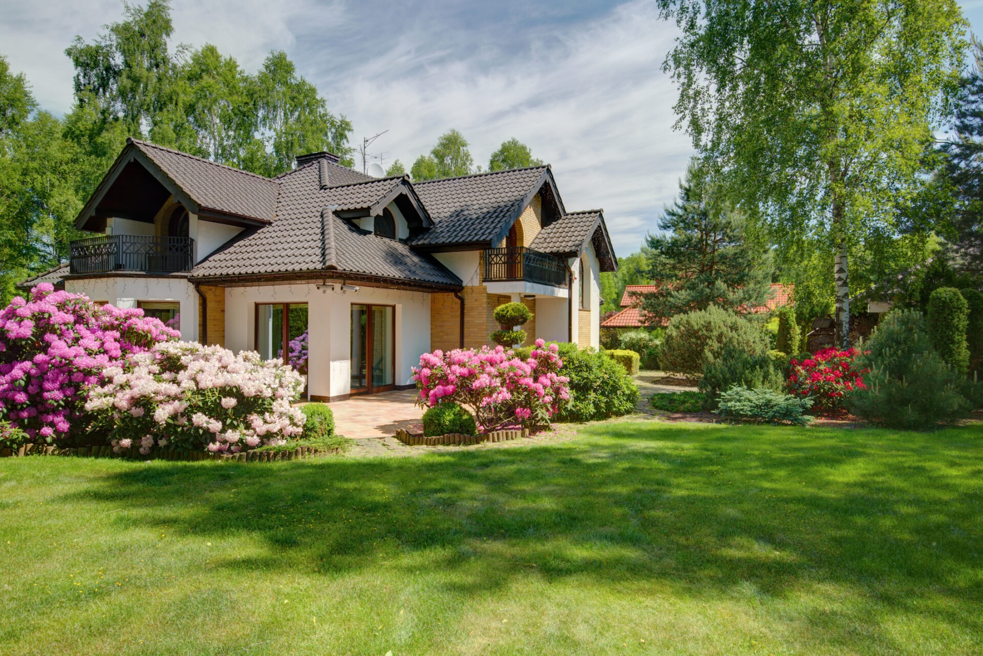 A suburban house with a dark roof is surrounded by vibrant blooming flowers, well-manicured lawn, and tall trees on a sunny day.