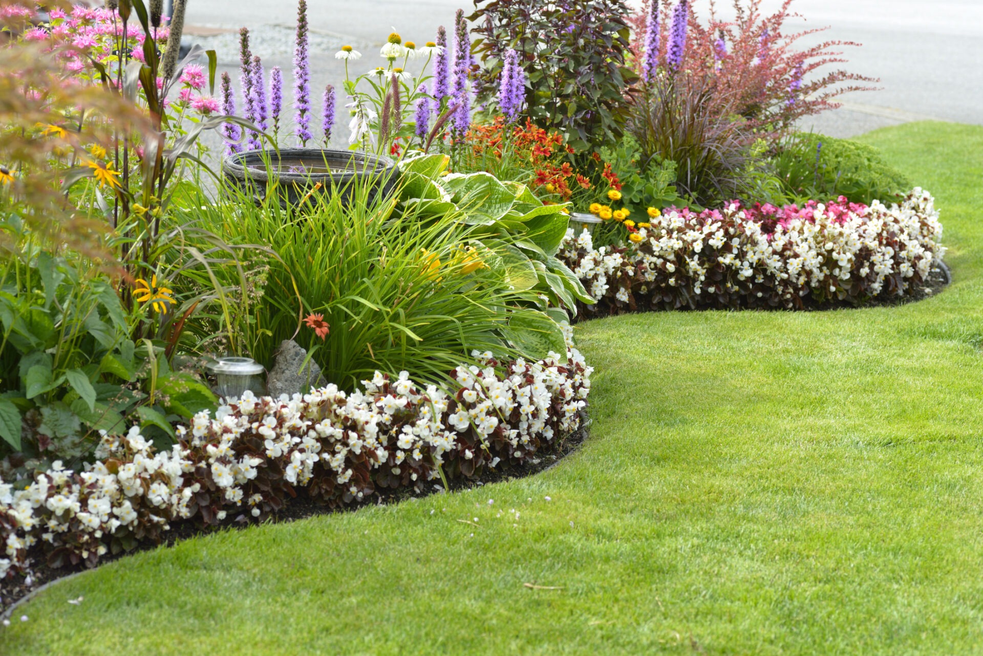 A neatly manicured garden with a variety of colorful flowers, lush green shrubs, and a curved pathway bordered by white blossoms.