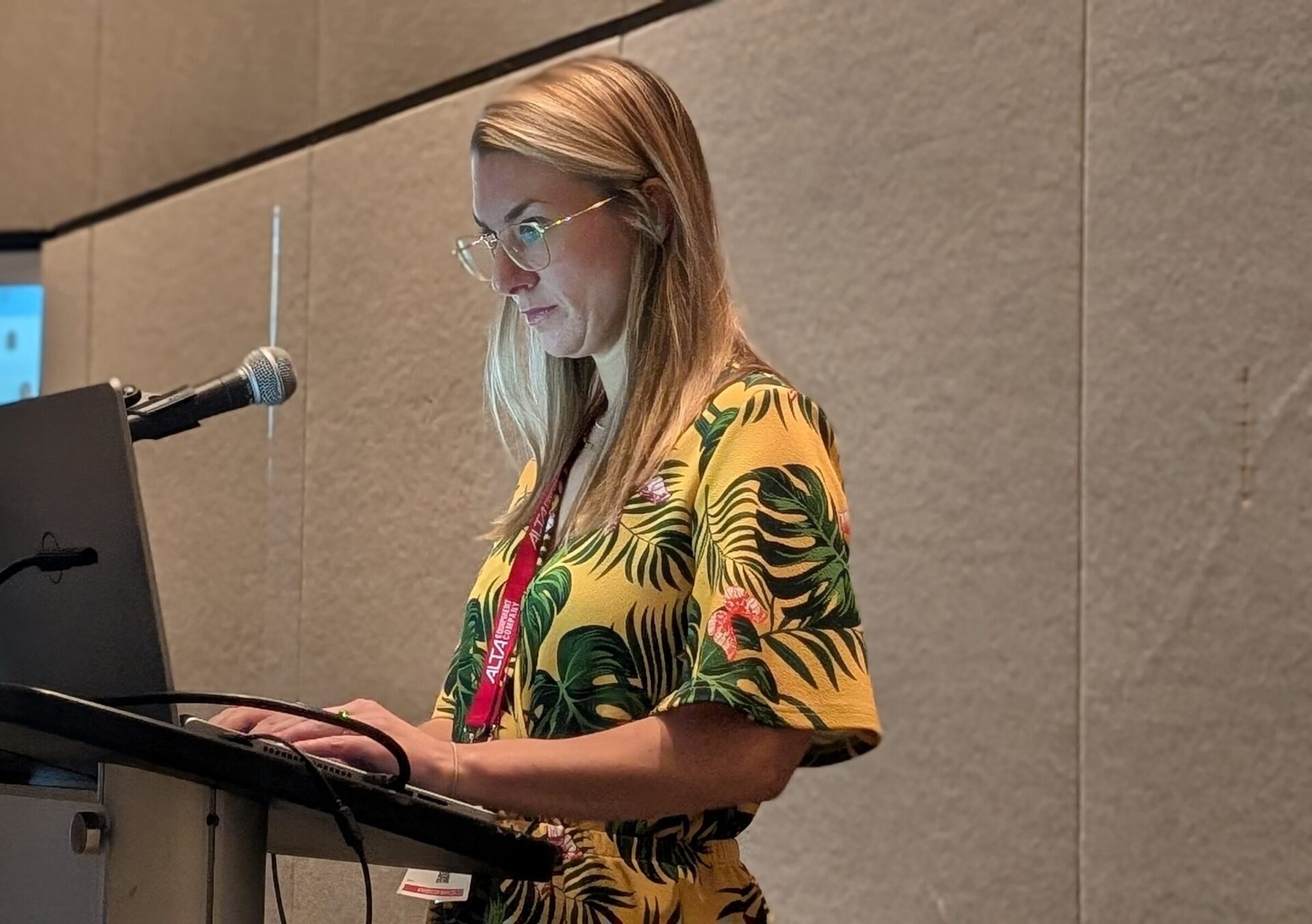 A person in a yellow floral shirt stands at a podium with a laptop and microphone, concentrating on a presentation in a modern conference room.