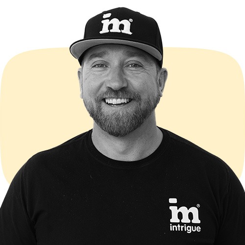 A person in a black shirt and hat, both with the "intrigue" logo, smiles against a beige background with a rounded rectangle shape.