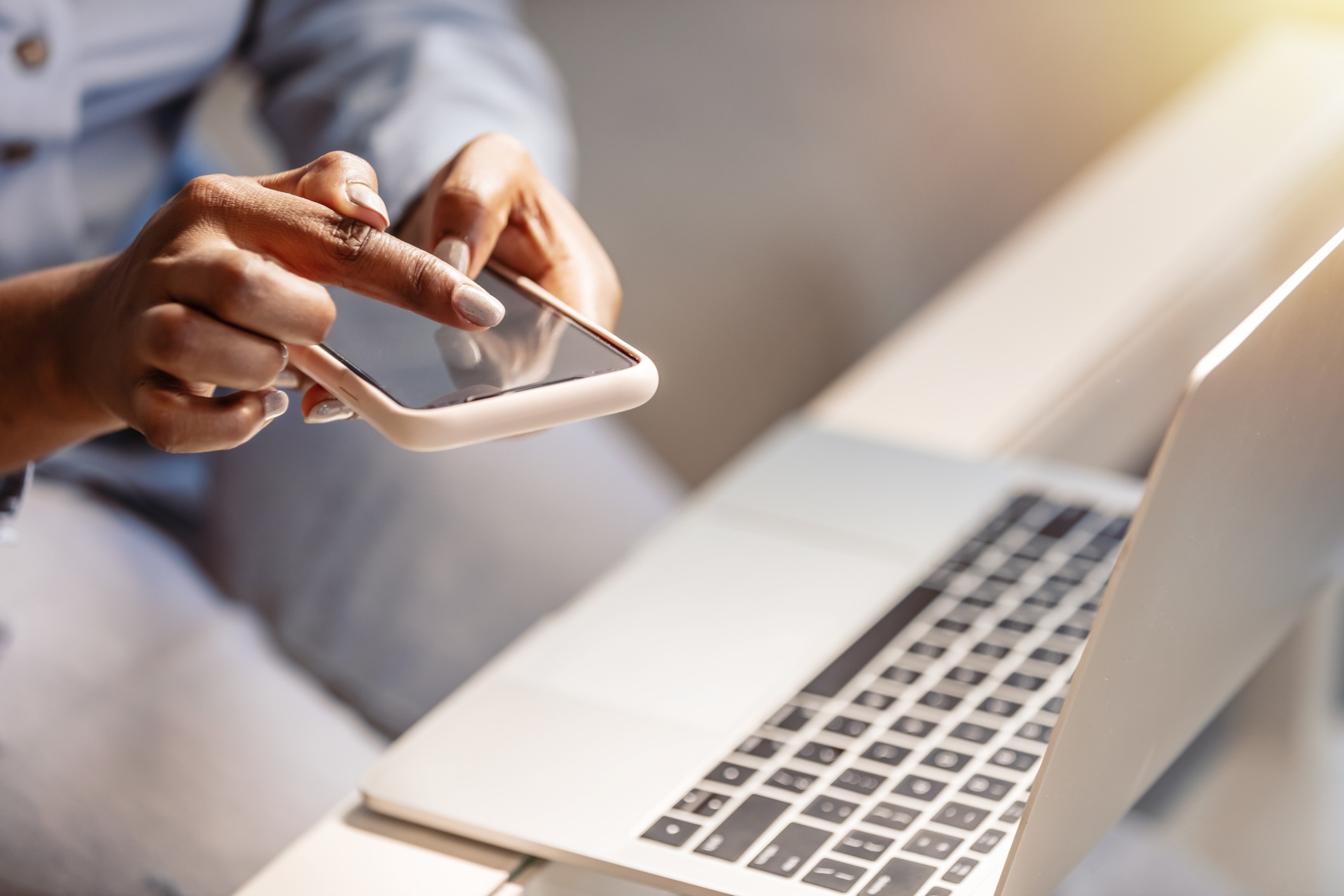 A person using a smartphone next to an open laptop, presumably working or browsing. The setting appears to be a well-lit indoor environment.