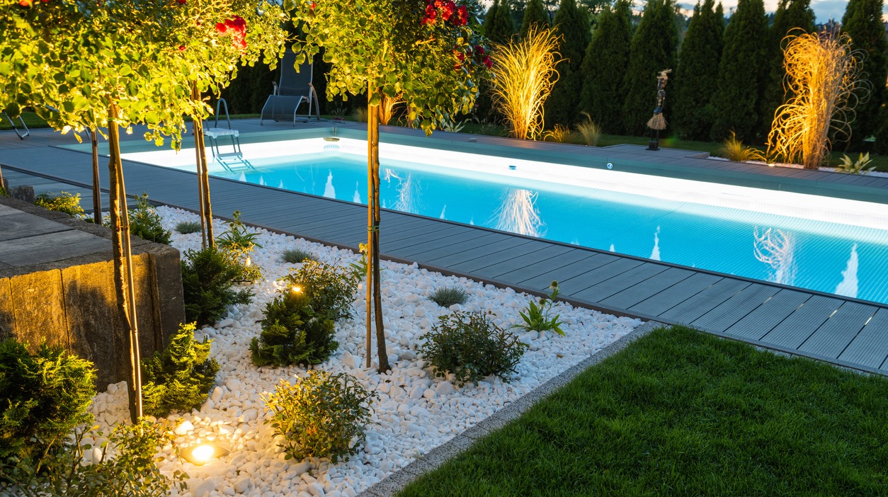 A serene backyard pool at dusk, surrounded by well-lit landscaping, decorative plants, and trees, creating a peaceful and inviting ambiance.