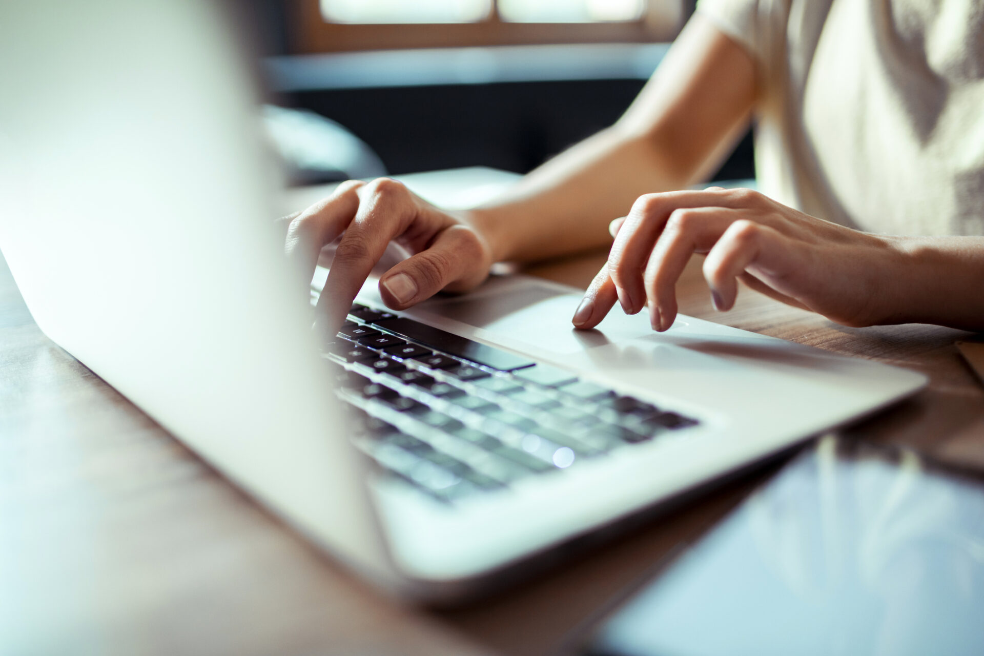 A person typing on a laptop keyboard with natural light coming through a window in the background. Smartphone visible on the table.