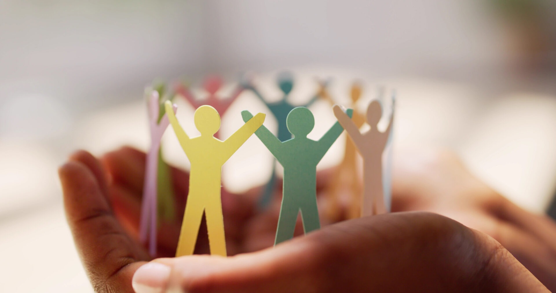 A person holds a paper chain of multicolored human figures, symbolizing unity and community. The background is soft and out-of-focus.