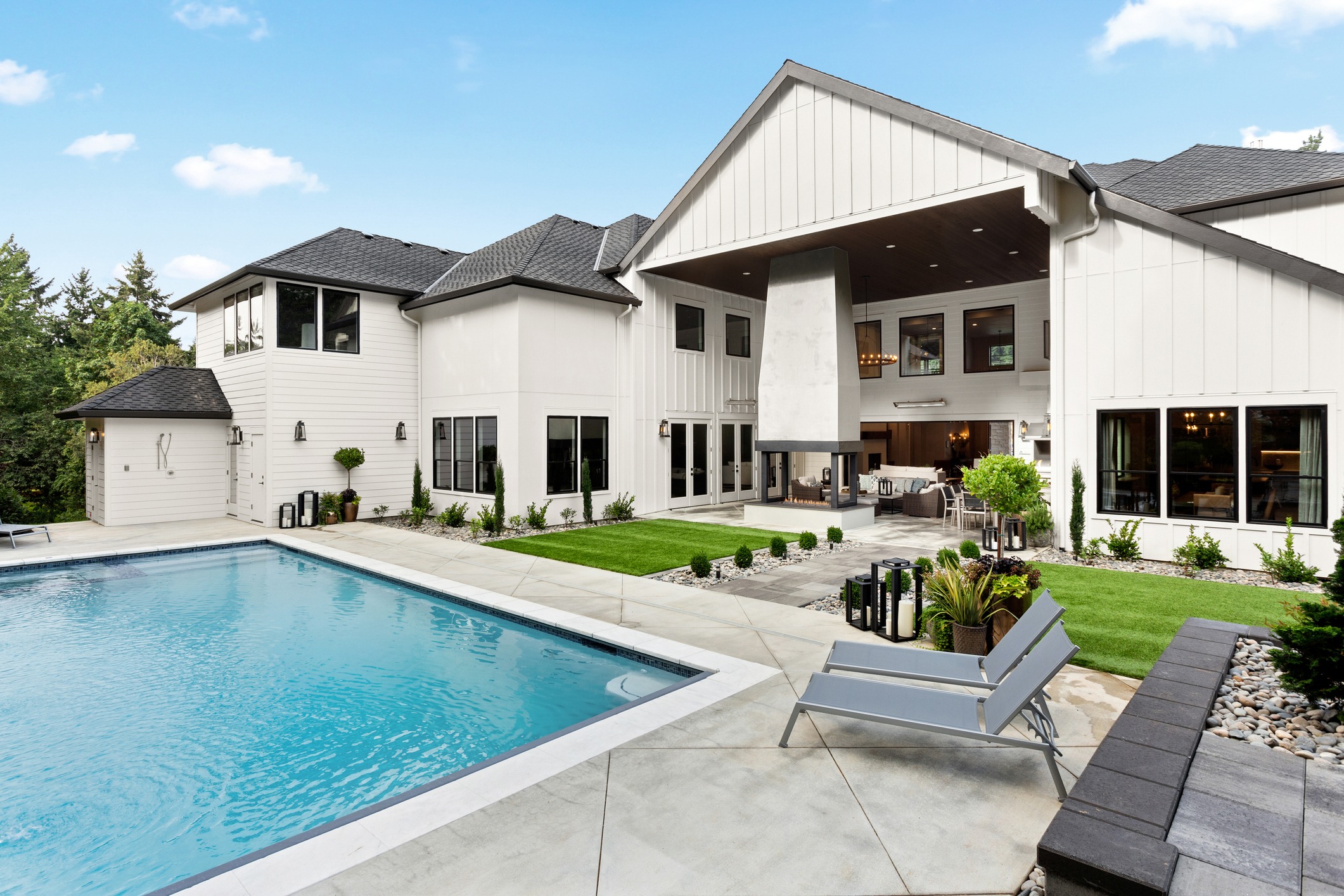 Modern two-story house with a swimming pool, outdoor seating area, and a covered patio with a fireplace, surrounded by landscaped greenery and trees.