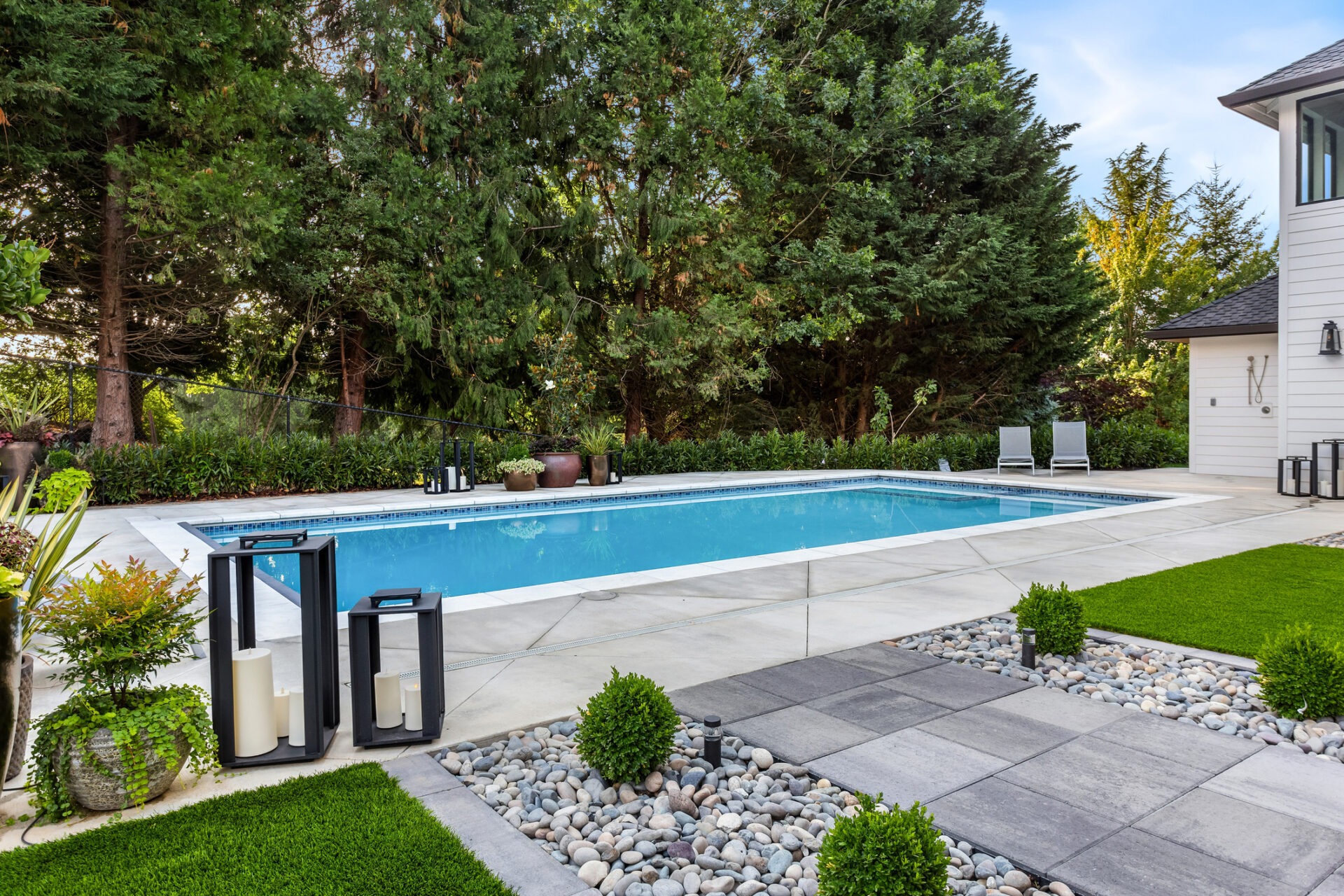 A modern backyard with a swimming pool, patio furniture, lanterns, and lush greenery. The area is neatly landscaped with stones and plants.