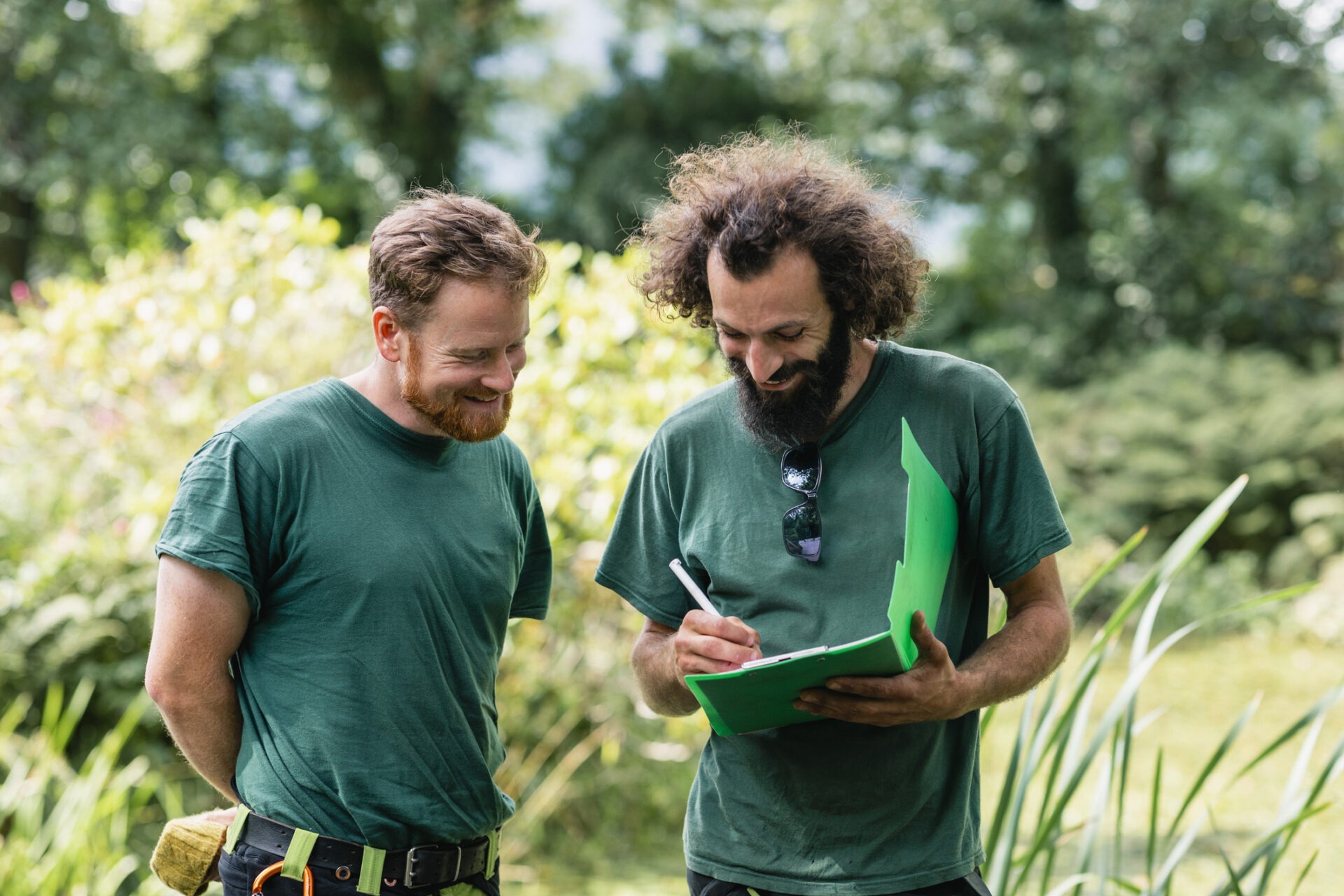 Two people in green shirts converse outdoors, one writes on a clipboard. Lush greenery surrounds them, suggesting a forest or garden setting.