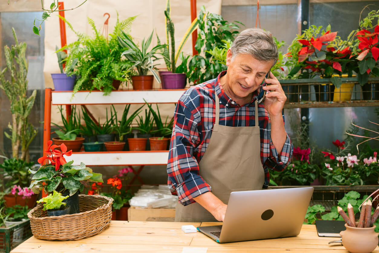 A person in an apron uses a laptop and phone in a greenhouse, surrounded by potted plants and flowers.