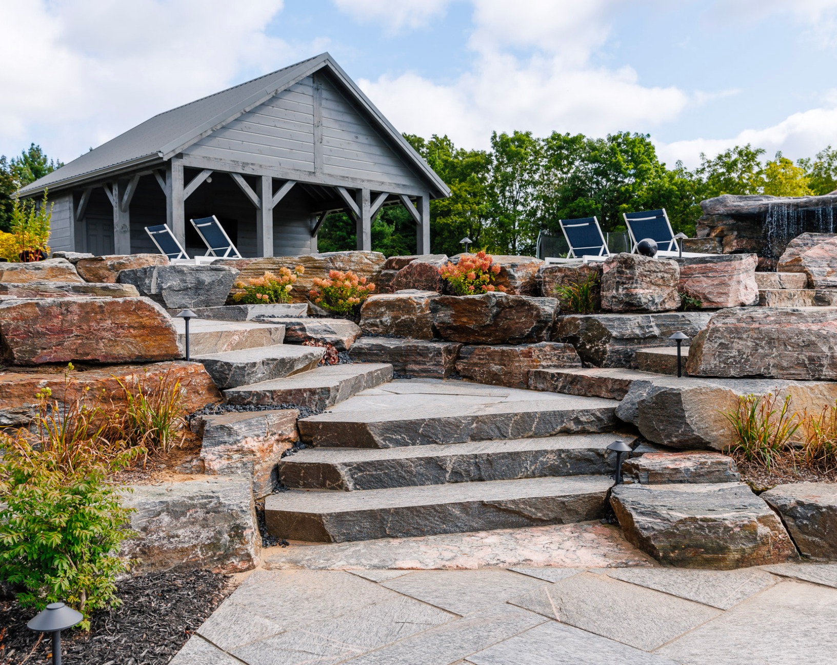 A rustic, wooden gazebo sits behind a stone terrace with steps and lounge chairs, surrounded by rocks, greenery, and a small waterfall.