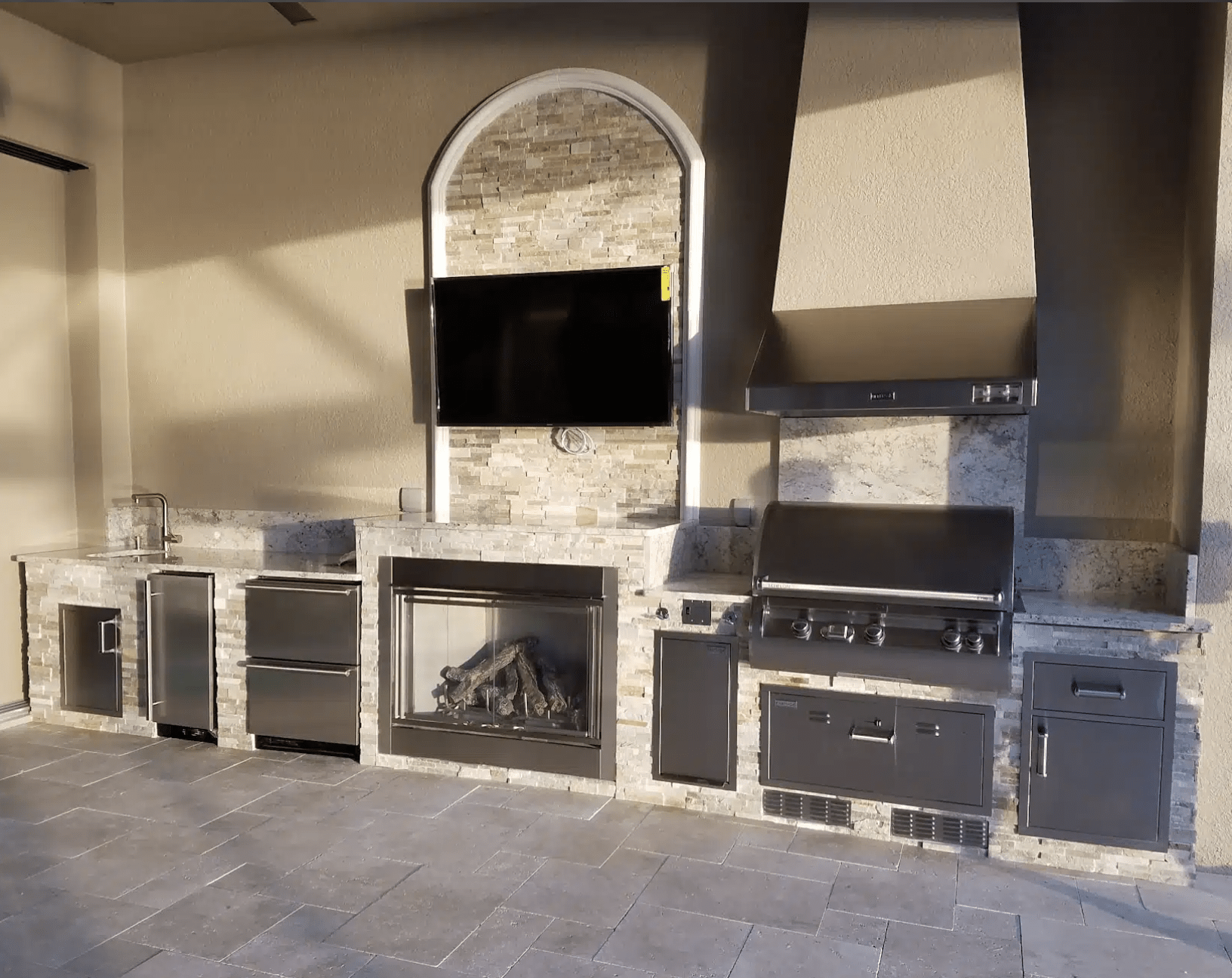 Outdoor kitchen with stainless steel appliances, stone countertops, built-in grill, fireplace, sink, and mounted TV on a tiled patio area. Minimalist and modern design.