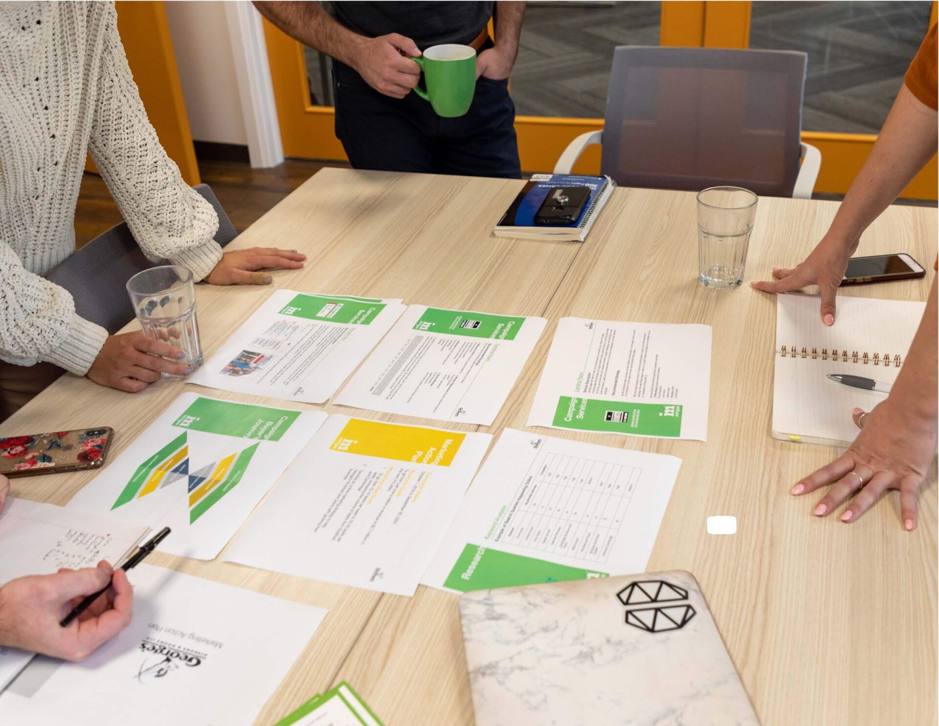 Four people collaborate around a table with documents and notebooks, engaged in a discussion or meeting. The focus is on planning.