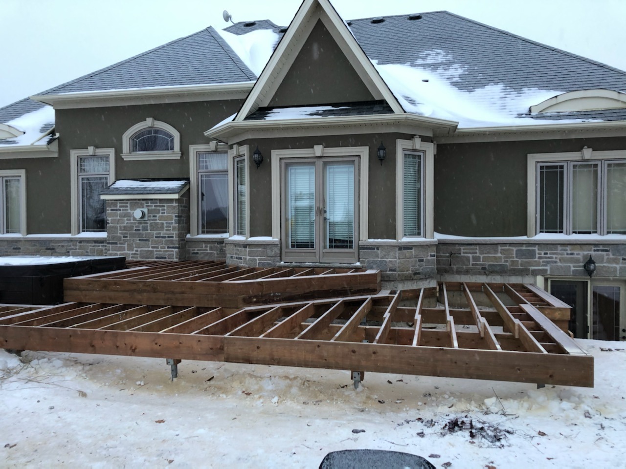 A snow-covered house with a wooden deck under construction in the yard. The green house has multiple windows and a gray stone exterior.