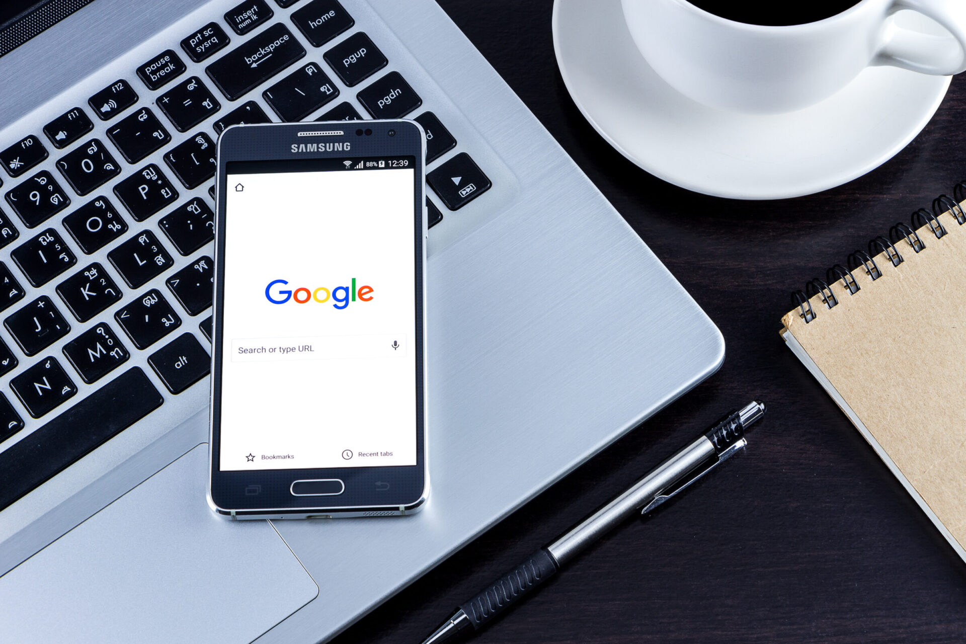 A Samsung phone displays Google search screen, placed on a laptop keyboard, accompanied by a coffee cup, pen, and notebook on a dark table.