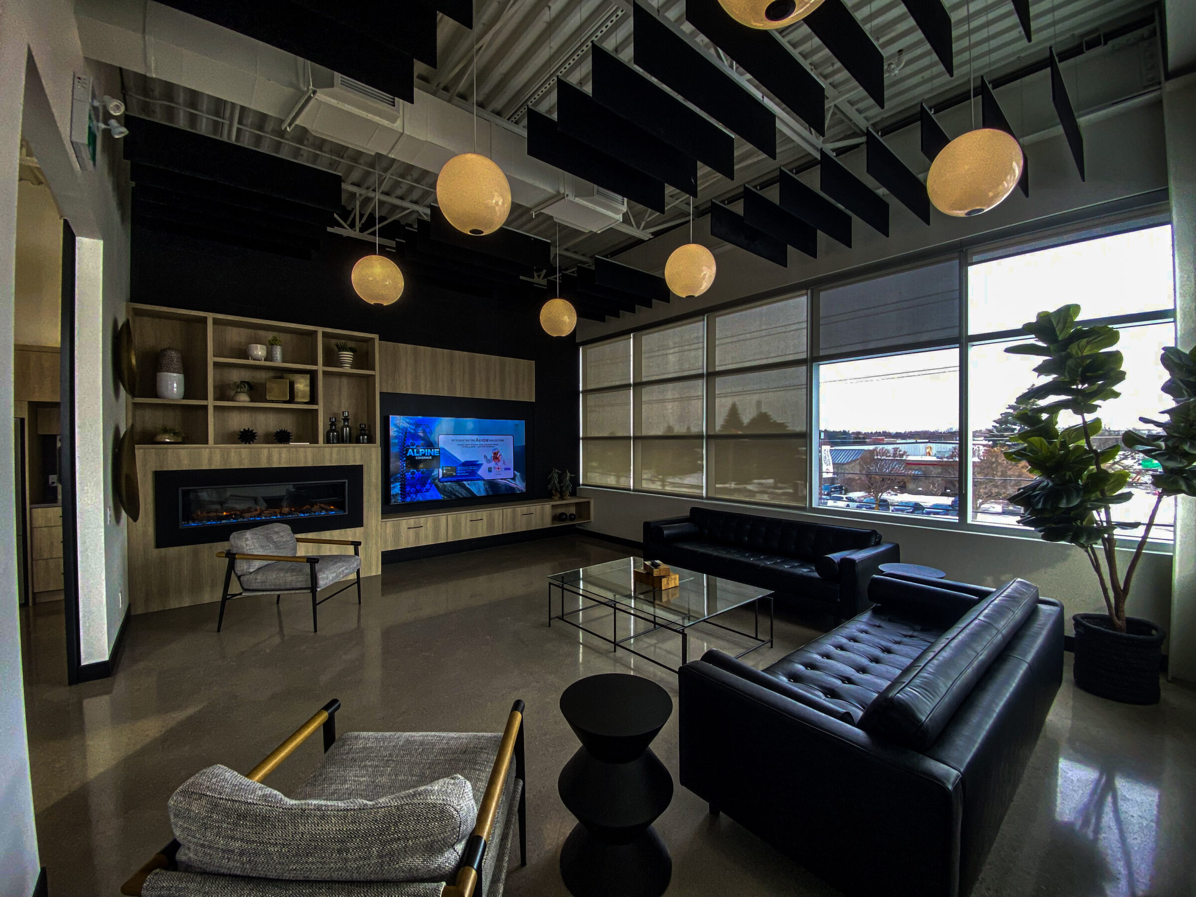 A modern lounge with leather sofas, a large TV, hanging lights, and big windows showing an urban landscape. Display shelves and plants add decor.