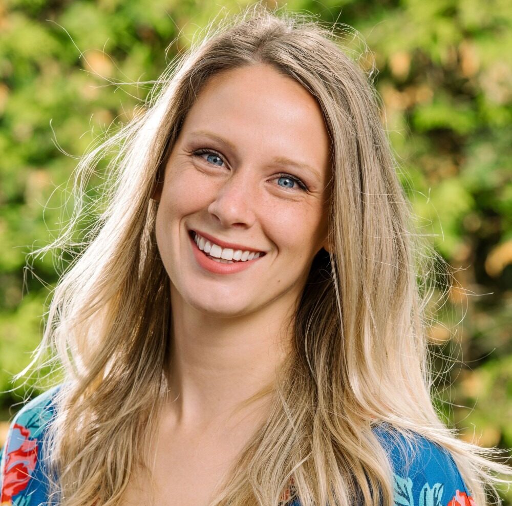 Smiling woman with long blonde hair wearing a blue floral blouse, standing outdoors with a green, leafy background.
