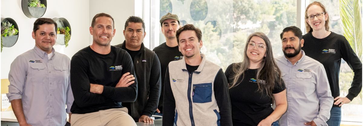 A team of eight professionals posing together in a modern office space, wearing branded shirts with the company logo. They are smiling and standing in a relaxed, collaborative pose.