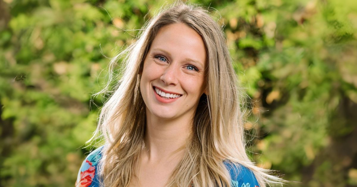 Smiling woman with long blonde hair wearing a blue floral blouse, standing outdoors with a green, leafy background.
