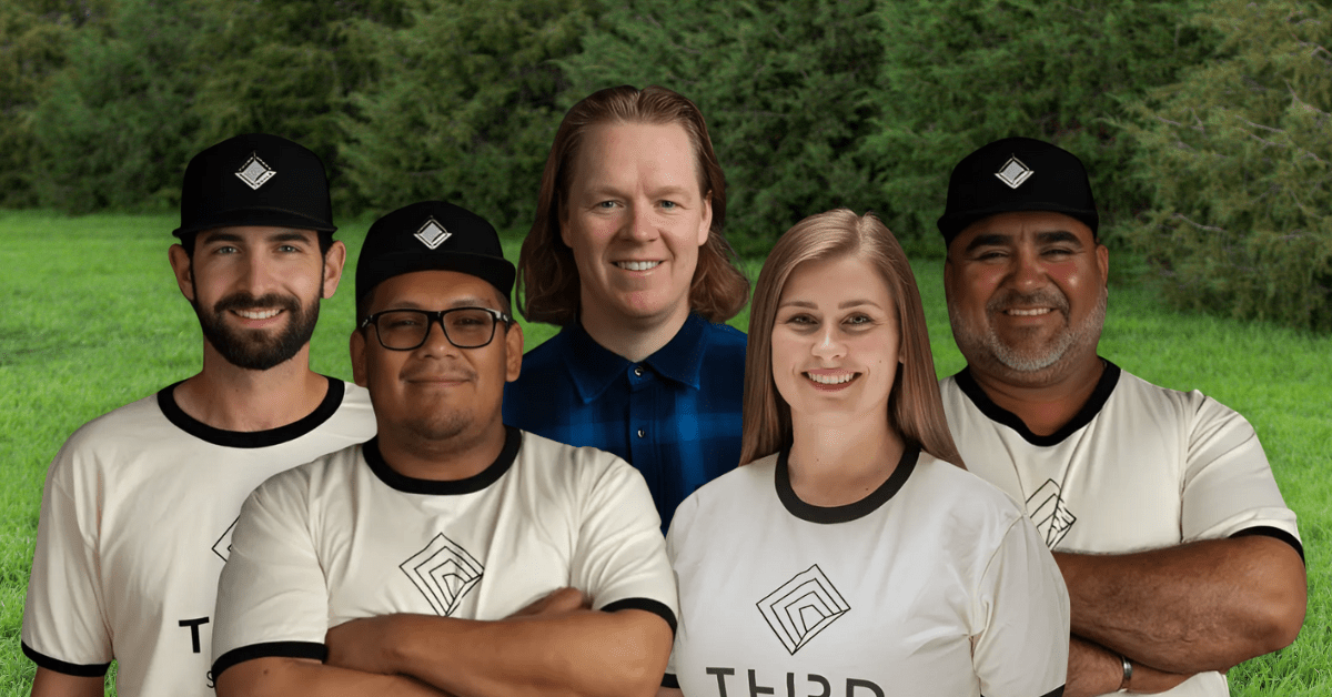 Group photo of the Third Space landscaping team standing outdoors. The team includes three men wearing branded Third Space shirts and black hats, a woman smiling in the center-right, and another man in a plaid shirt at the center. Trees and greenery are visible in the background.