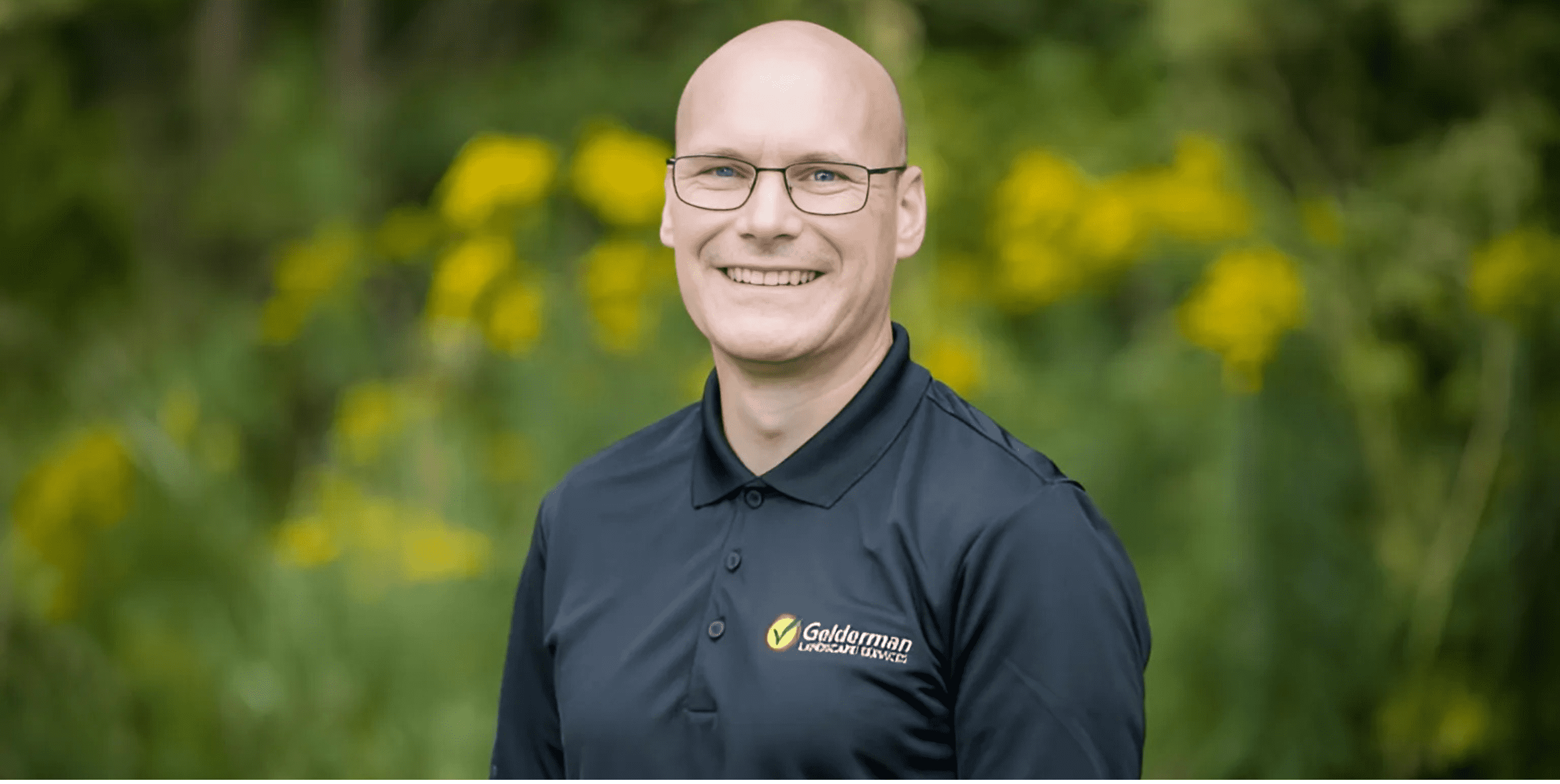 Smiling man wearing glasses and a Golderman Landscape Design polo shirt, standing outdoors with yellow flowers and greenery in the background