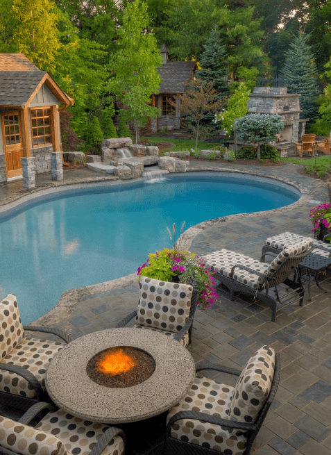 Luxurious backyard featuring a freeform in-ground pool surrounded by stone pavers, cushioned patio furniture with polka dot upholstery, a round fire pit table, lush landscaping, and a stone outdoor fireplace. A wood and stone pool house is visible in the background, framed by mature trees and colorful flowers.