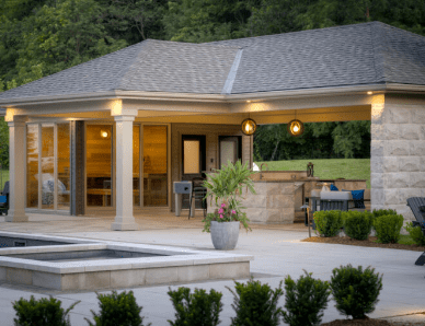 Modern backyard pavilion with a gable roof, stone columns, and built-in outdoor kitchen, featuring pendant lighting, bar seating, and a nearby plunge pool. The surrounding landscape includes manicured shrubs, potted plants, and a forested backdrop.